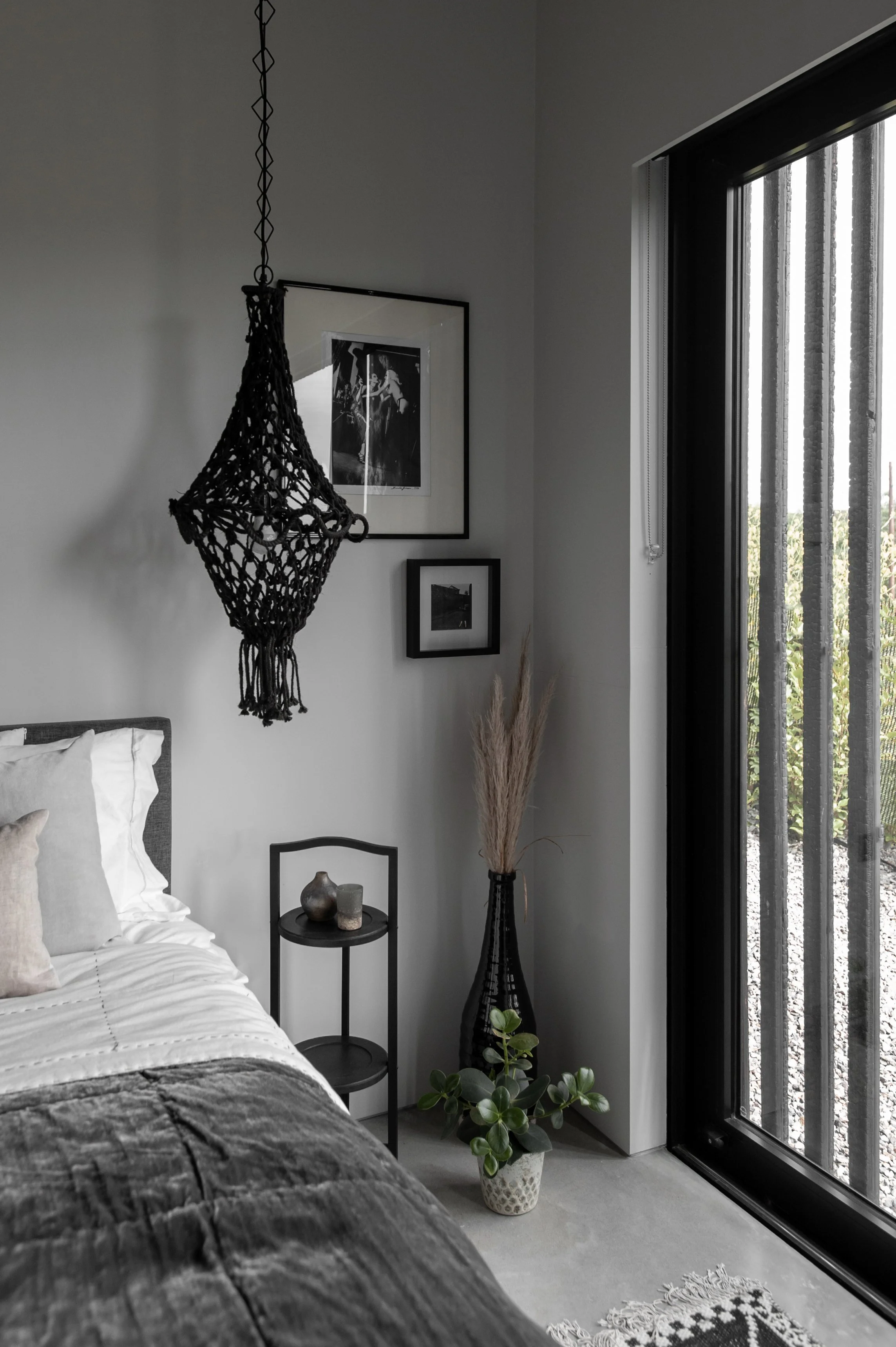 A gray bedroom corner with a bed, black nightstand with vases, large black vase with dried pampas grass, potted plant, black framed photos on the wall, a black decorative hanging, and a sliding glass door with vertical blinds showing an outdoor view.