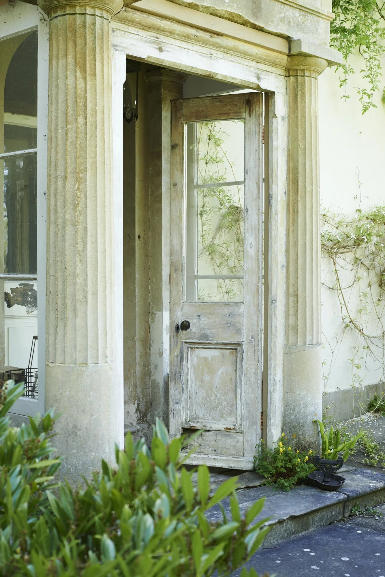Old weathered wooden door with glass pane, set in a stone archway, surrounded by greenery and plants, with a stone step at the entrance.