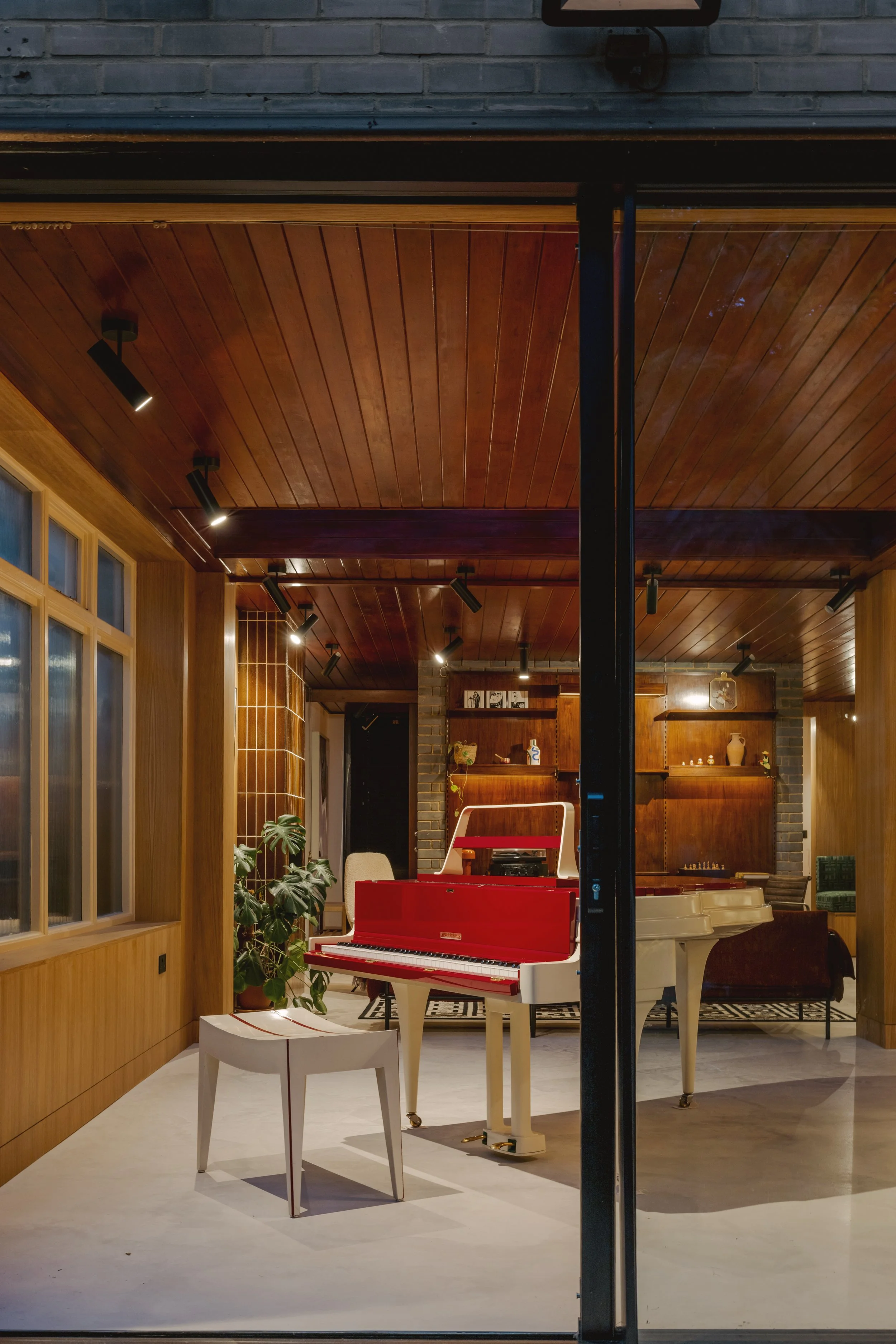 Interior of a music room with a red and white piano, a matching piano bench, wooden walls and ceiling, spotlights, and decorative items on wooden shelves.