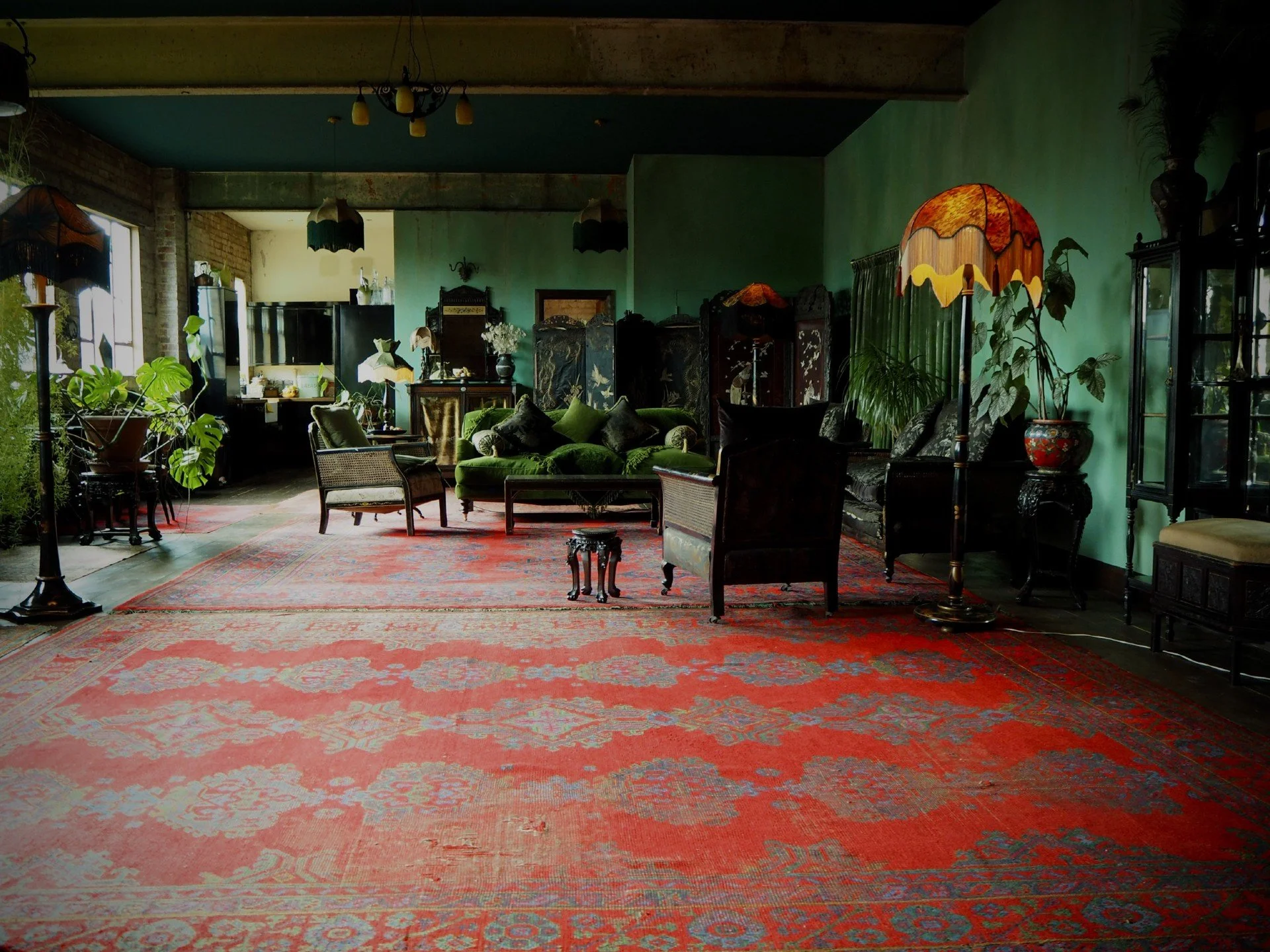 Living room with green walls, vintage furniture, potted plants, patterned rugs, and multiple lamps.