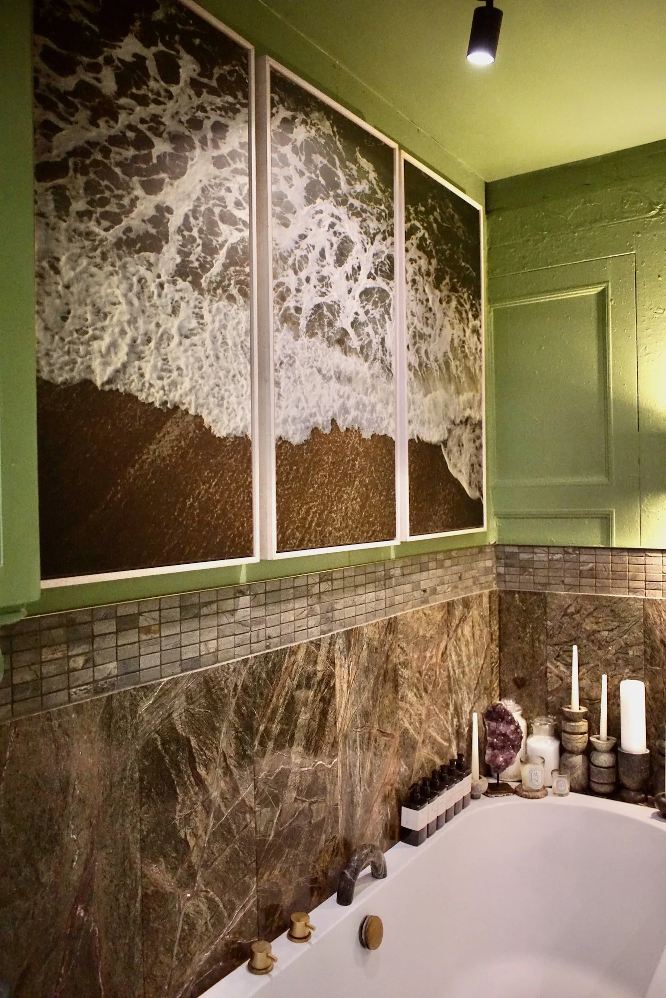 A bathroom wall decorated with a triptych of ocean wave art, with a green painted ceiling and a corner of a bathtub filled with candles and decor items.