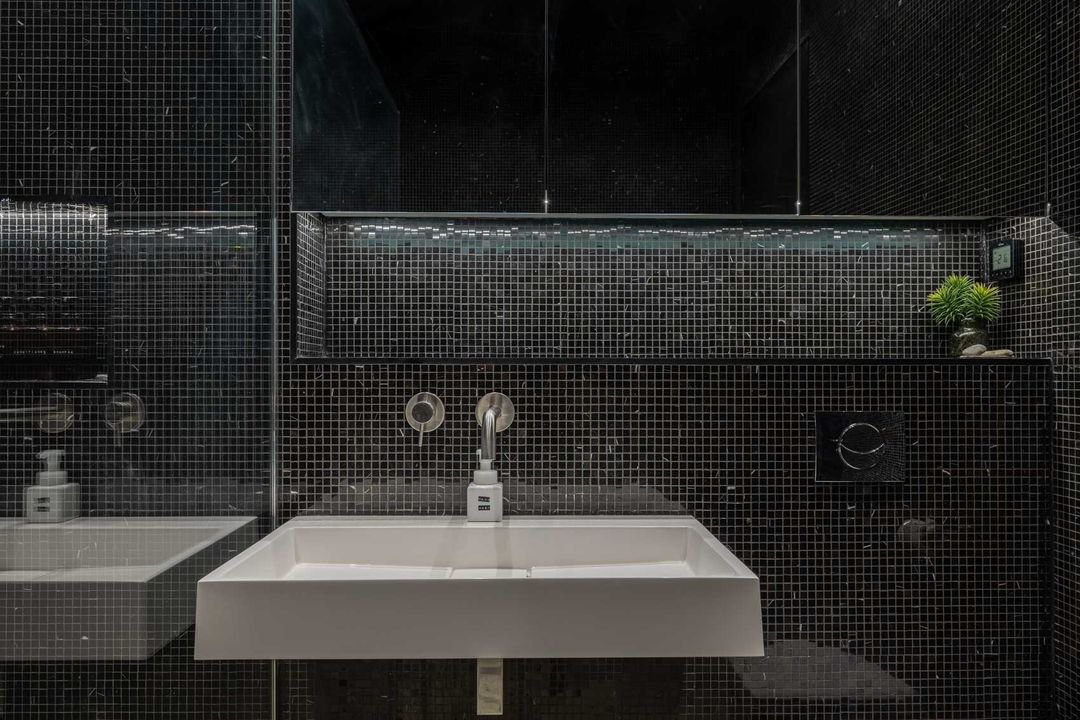 Modern bathroom with black tiled walls, a white rectangular sink, a small plant on a shelf, a soap dispenser, and a mirror.