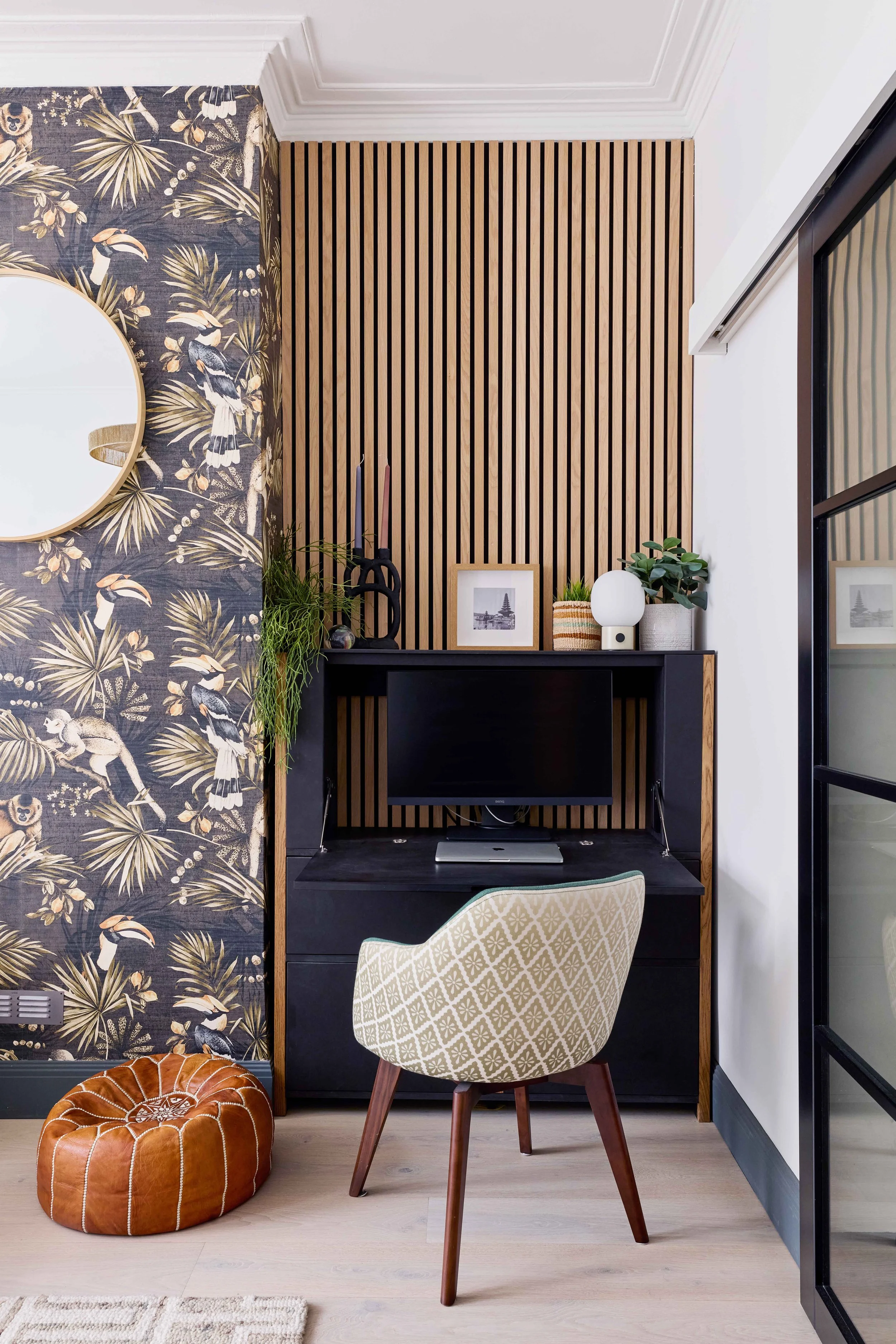 A home office with a black desk, a computer, decorative vases, and framed pictures, against a wooden slat wall and a wallpapered wall with botanical and toucan motifs, with a beige patterned chair and a brown leather pouf.