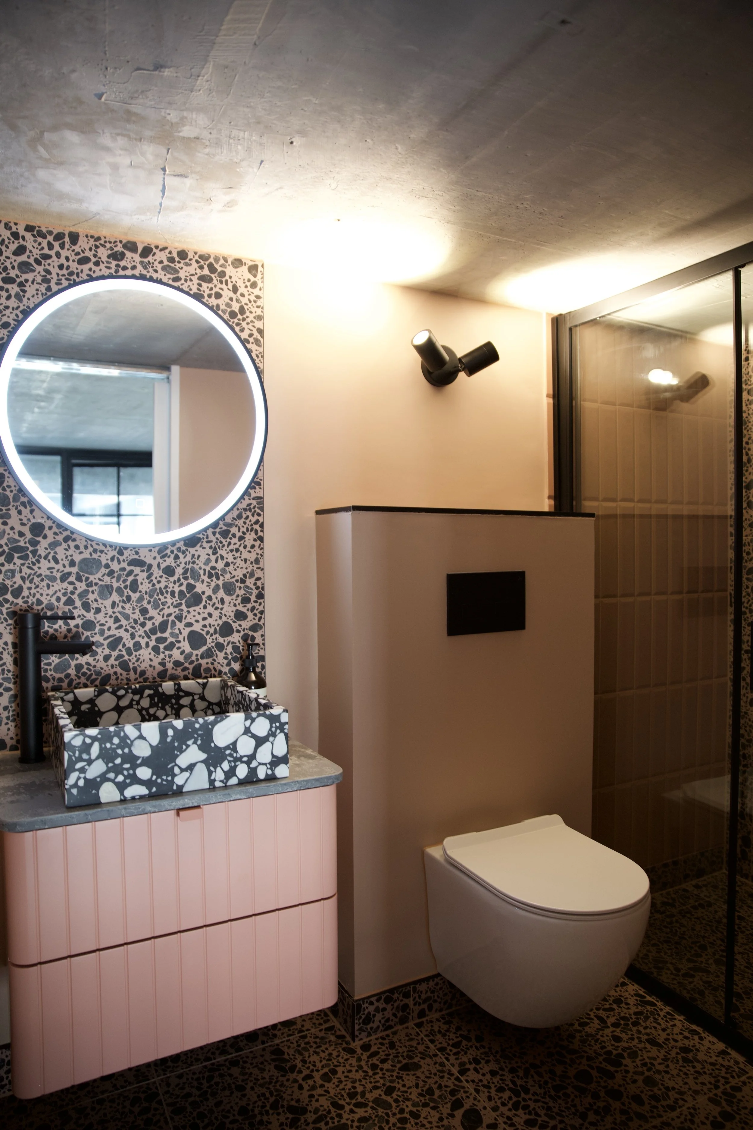 Modern bathroom featuring a round illuminated mirror, patterned black and white sink, black faucet, pink vanity, wall-mounted toilet, and a black shower enclosure with beige tiled interior.