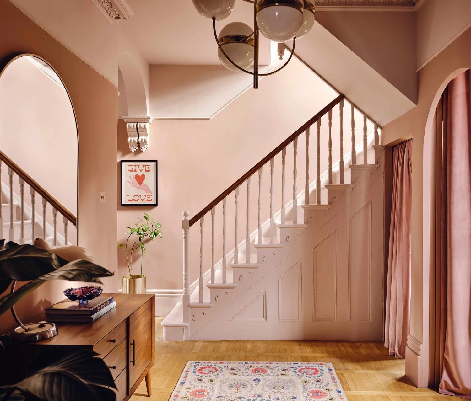 A cozy living room with a staircase and pink curtains. There is a mid-century wooden sideboard with books, a lamp, and a decorative bowl. A potted plant is beside a framed art print that says 'Give Love' with a heart and hands. A colorful rug is on t