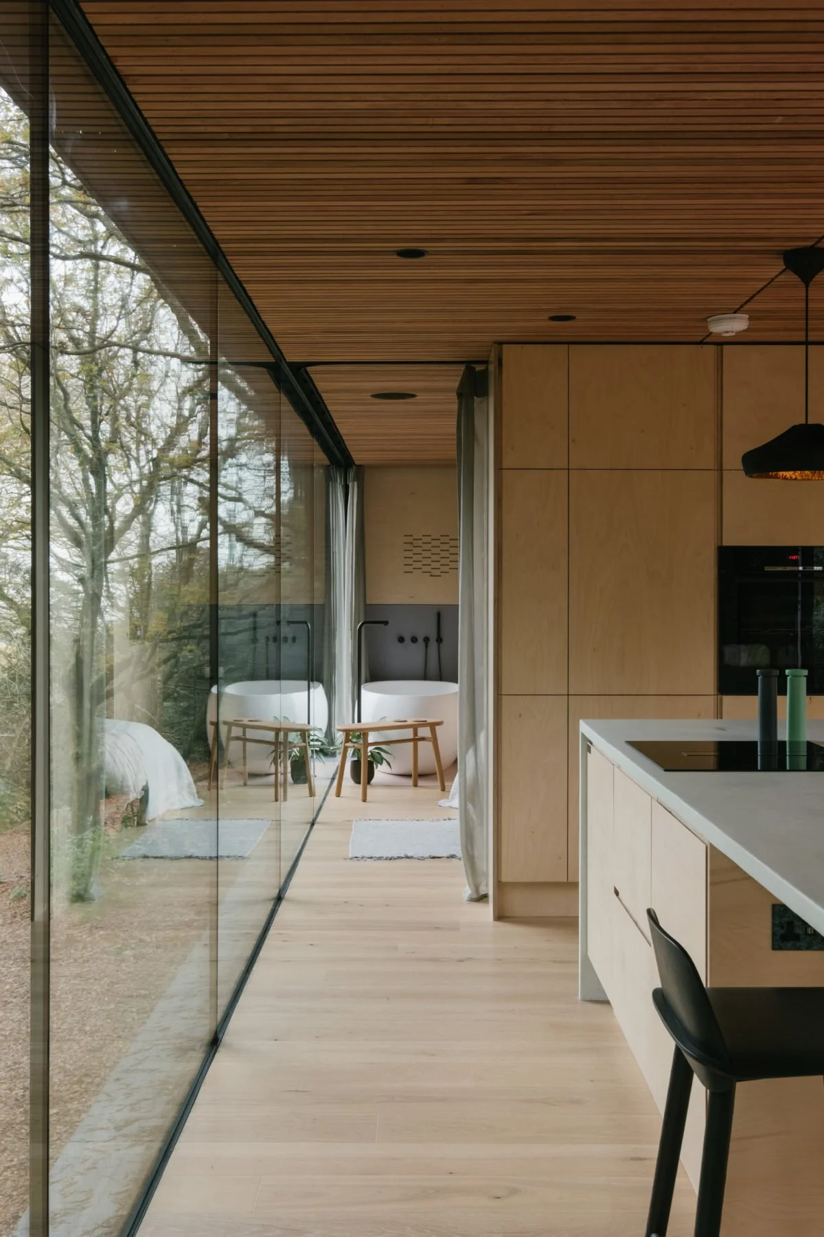 Modern kitchen with wooden ceiling and floor, glass wall overlooking outdoor trees, and a sitting area with two freestanding bathtubs and chairs.