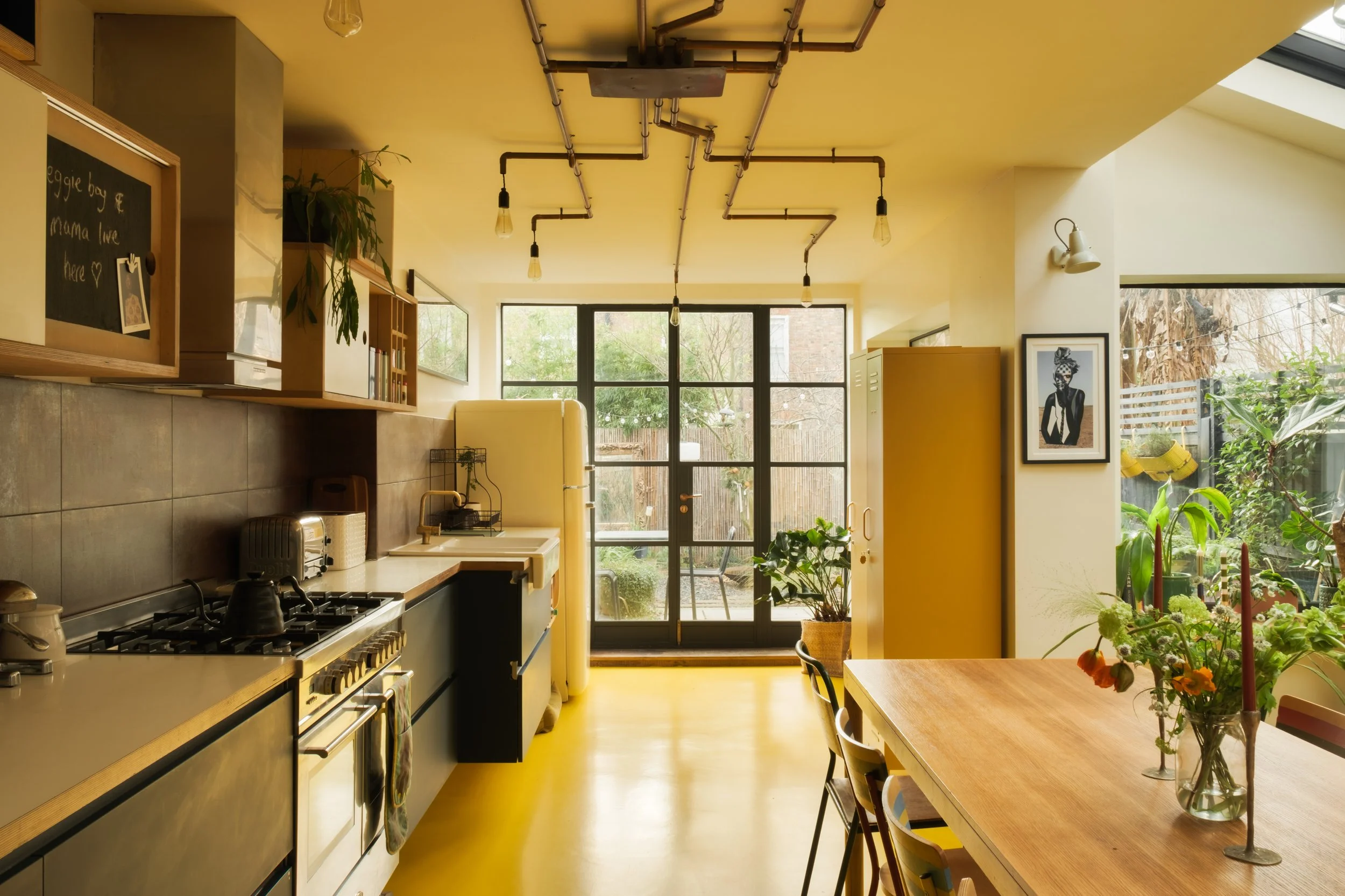 Bright kitchen with black-framed glass door leading to the outdoor garden, yellow walls and ceiling, wooden table with flowers, hanging lightbulbs, potted plants, and a framed artwork.