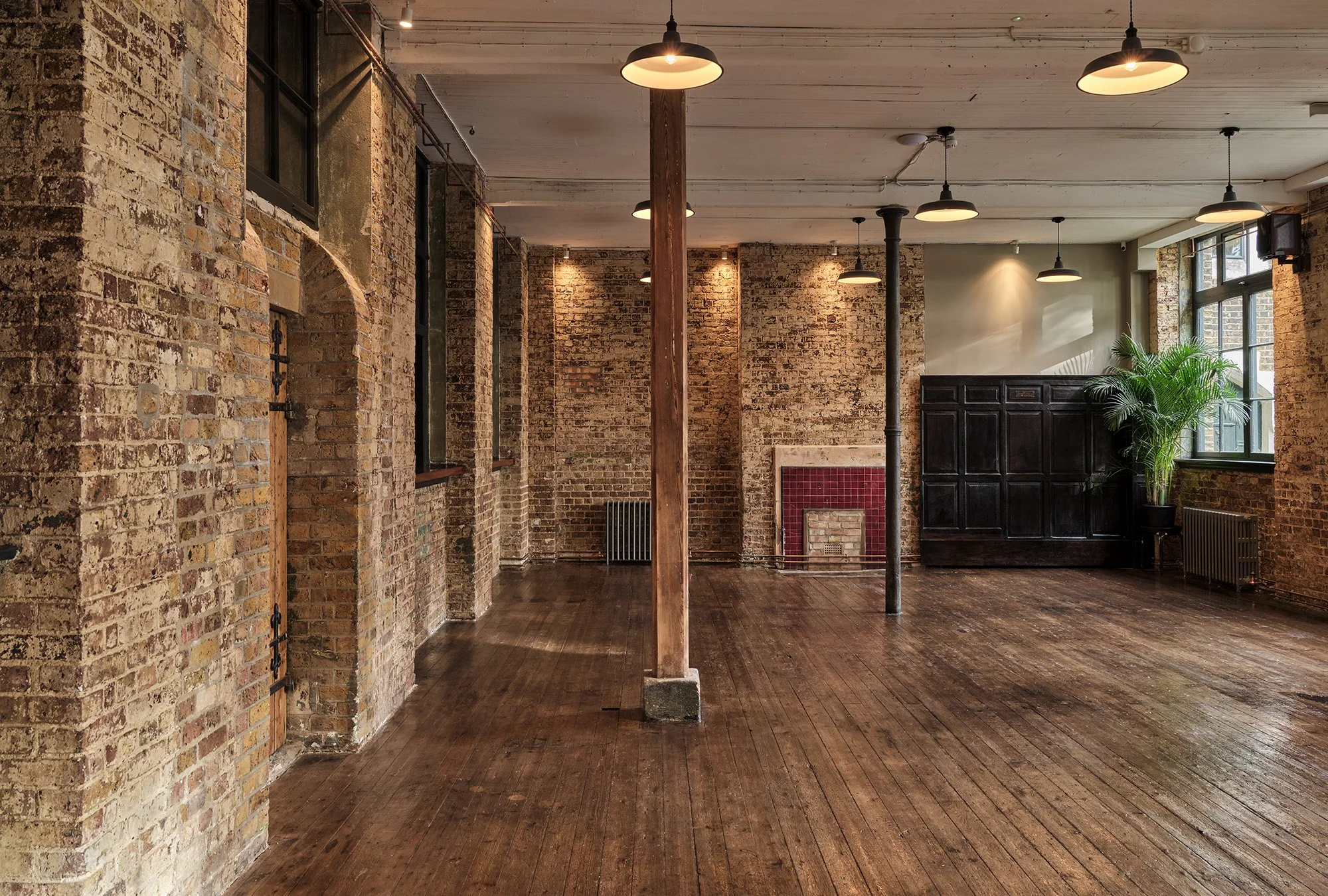 Empty industrial-style room with exposed brick walls, wooden floors, black ceiling lights, large windows, a fireplace with red tiles, and a potted plant.