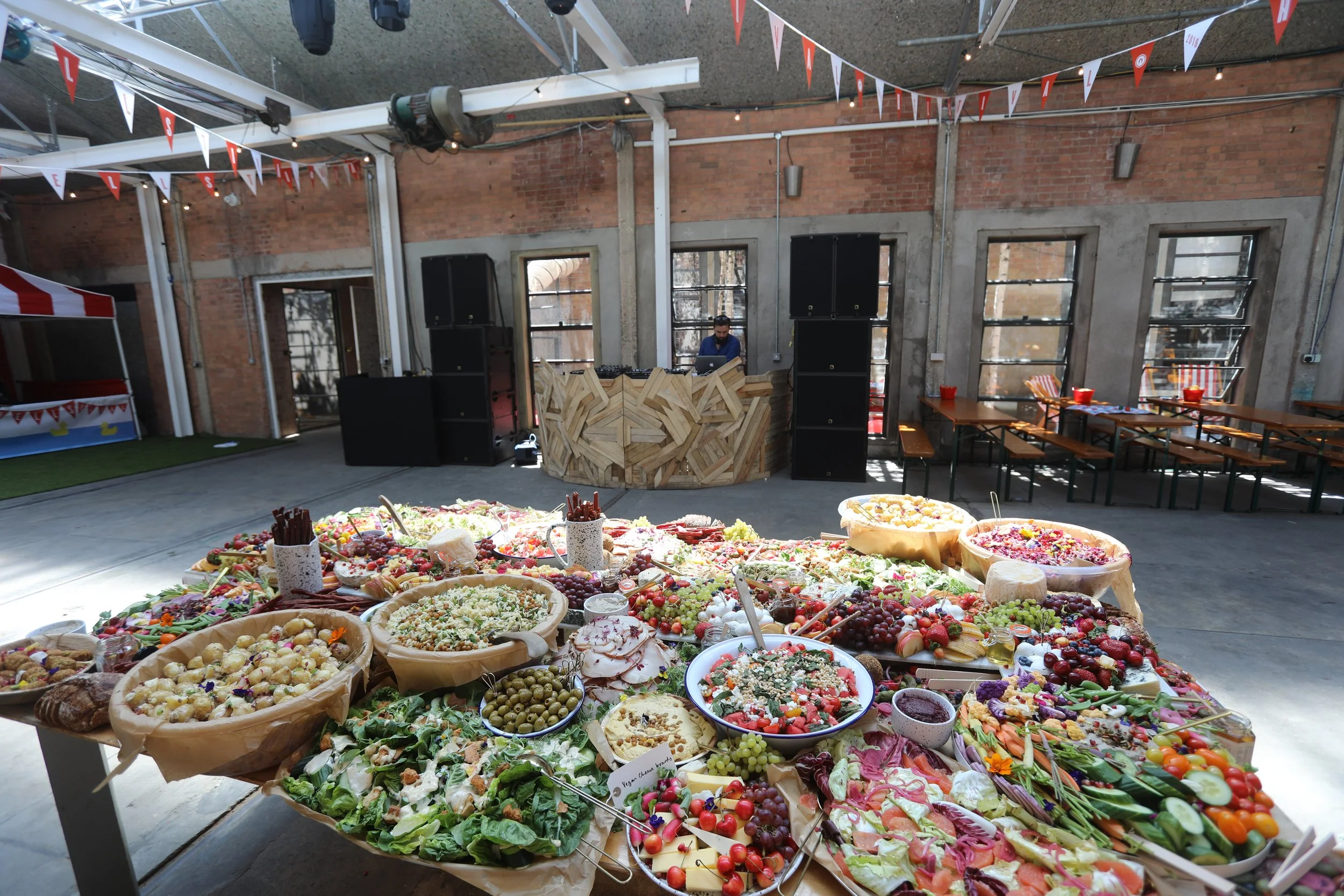 A large table filled with assorted fruits, salads, and snacks at an indoor event or party, with a DJ booth and seating area in the background.