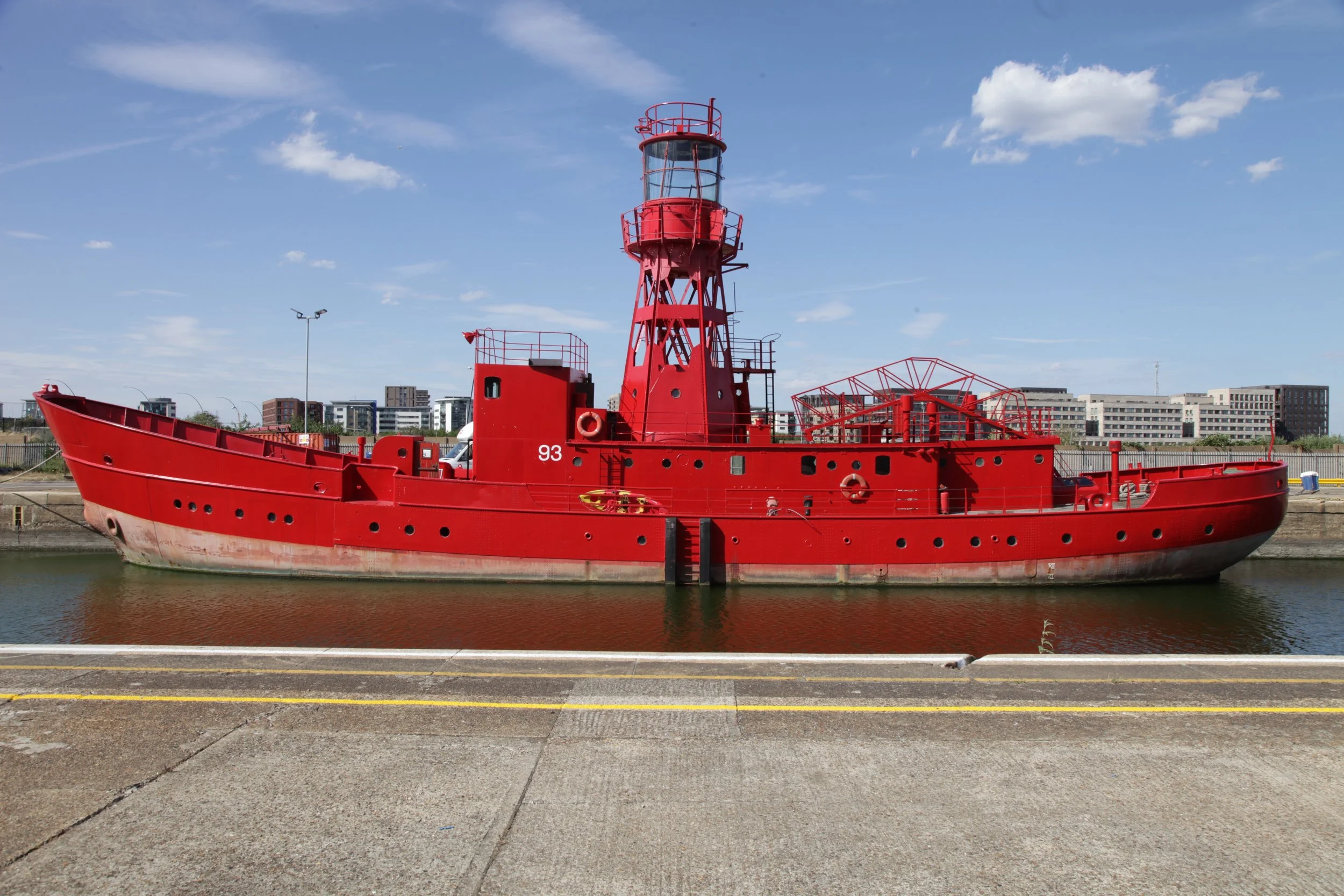 Red lighthouse boat on water with city buildings in background under blue sky with clouds.
