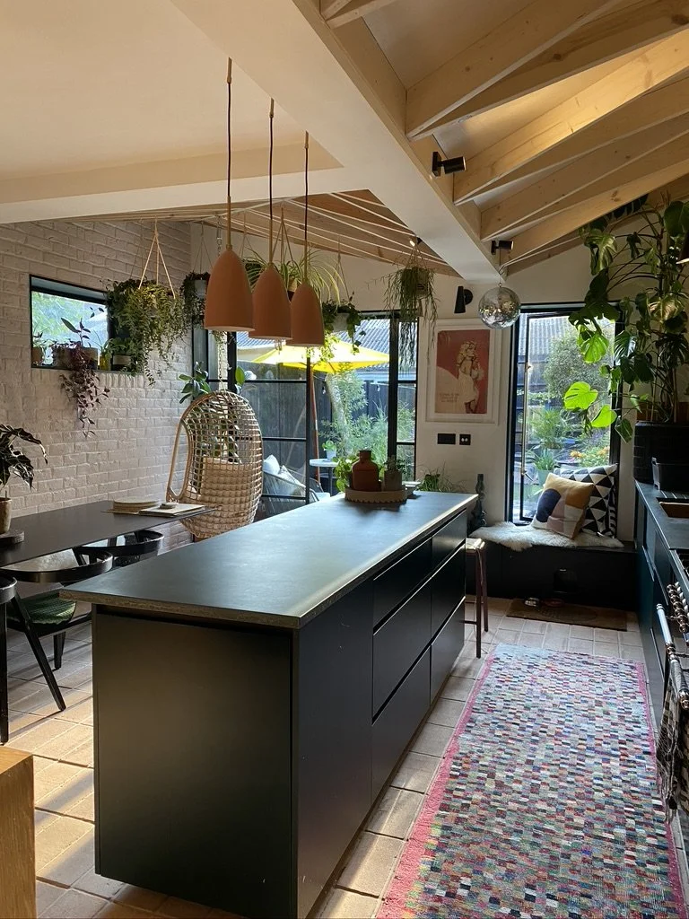 A cozy kitchen with a black island, hanging pendant lights, a woven hanging chair, green plants, and a windowed seating nook with decorated pillows.