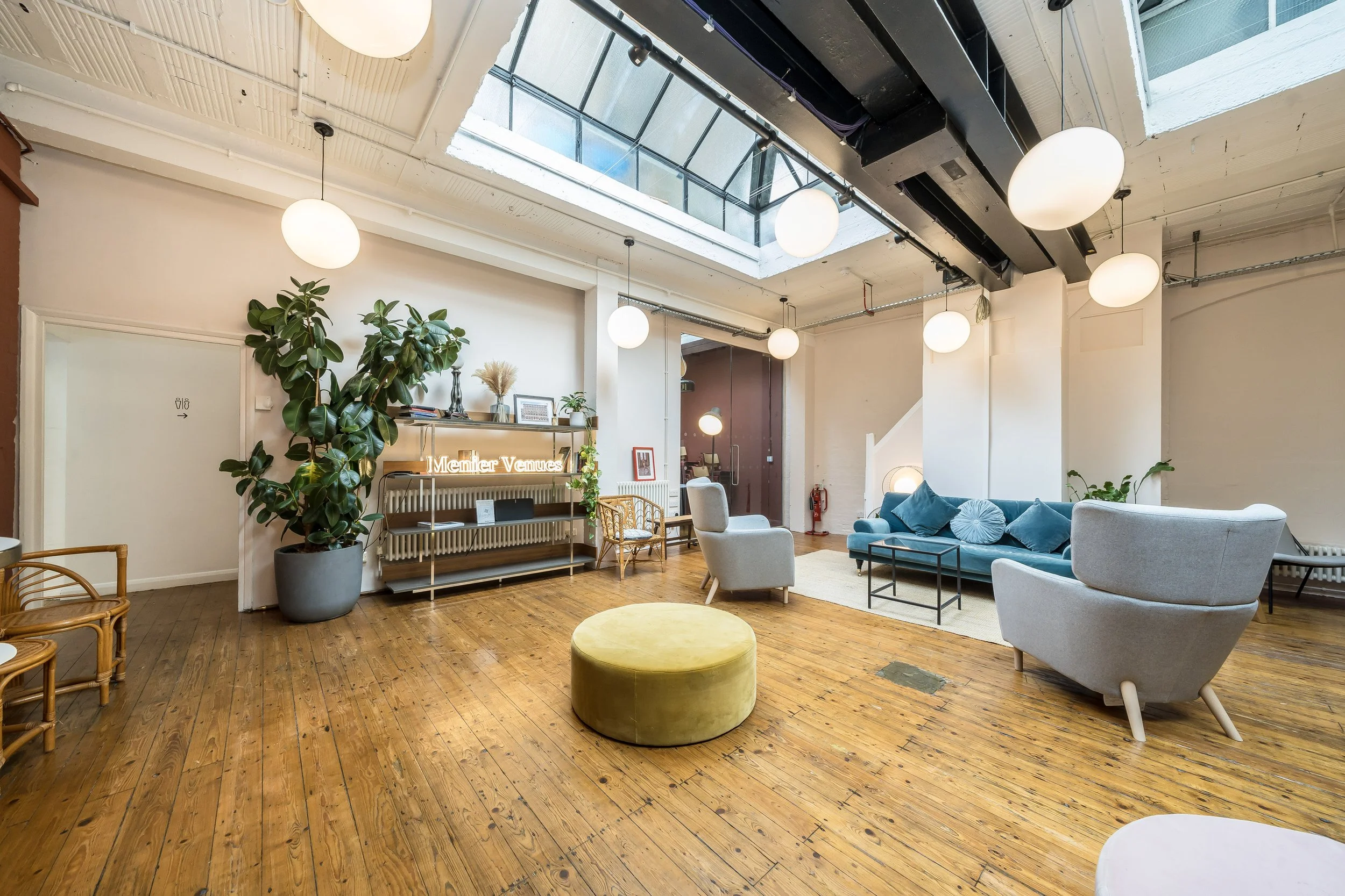 A spacious, modern living room with wooden floors, white walls, and a large skylight ceiling with circular light fixtures. The room features a blue sofa, light gray armchairs, a yellow ottoman, and various plants. There's minimal decor, including a s