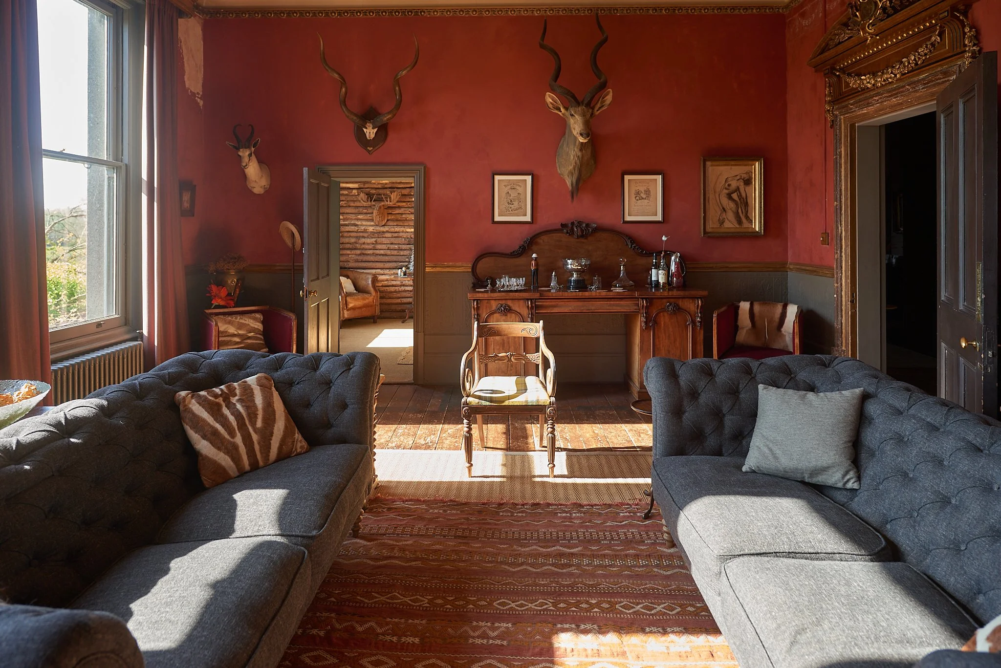 A richly decorated vintage living room with red and gray walls, two tufted gray sofas, and a wooden chair in the center. Decor includes animal horns and mounted animal heads on the walls, framed artwork, a wooden sideboard with silver cups and glassw