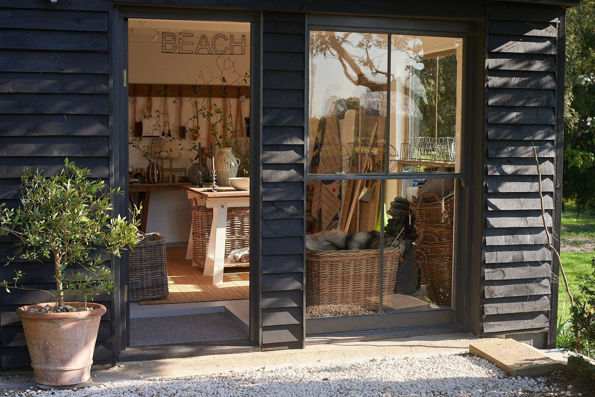 View of a small black wooden shop or studio with large window and door, inside are various home decor items, plants, baskets, and art supplies, with a sign that reads 'BEACH' visible inside.