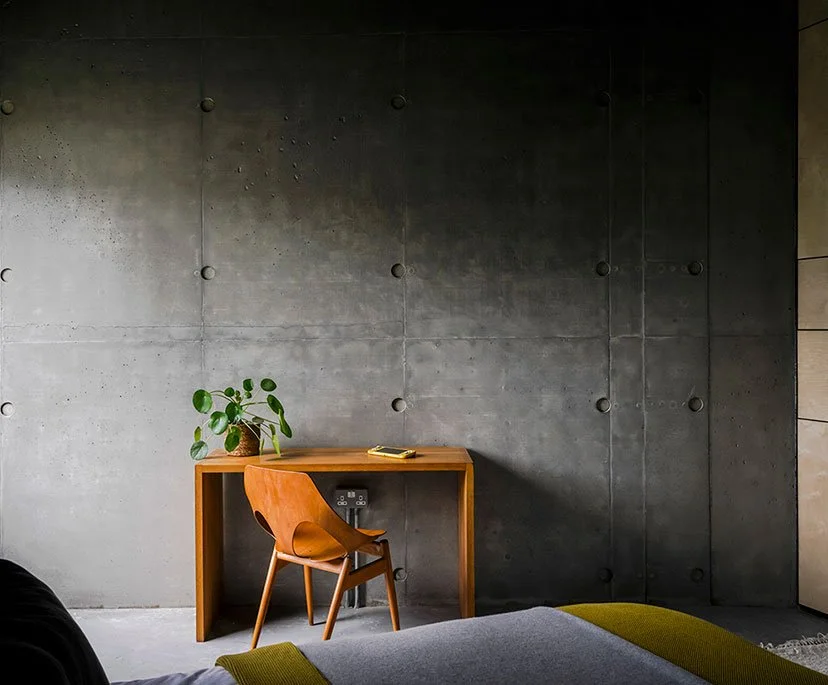 A minimalist workspace with a wooden desk, an orange chair, and a potted plant against a dark concrete wall.