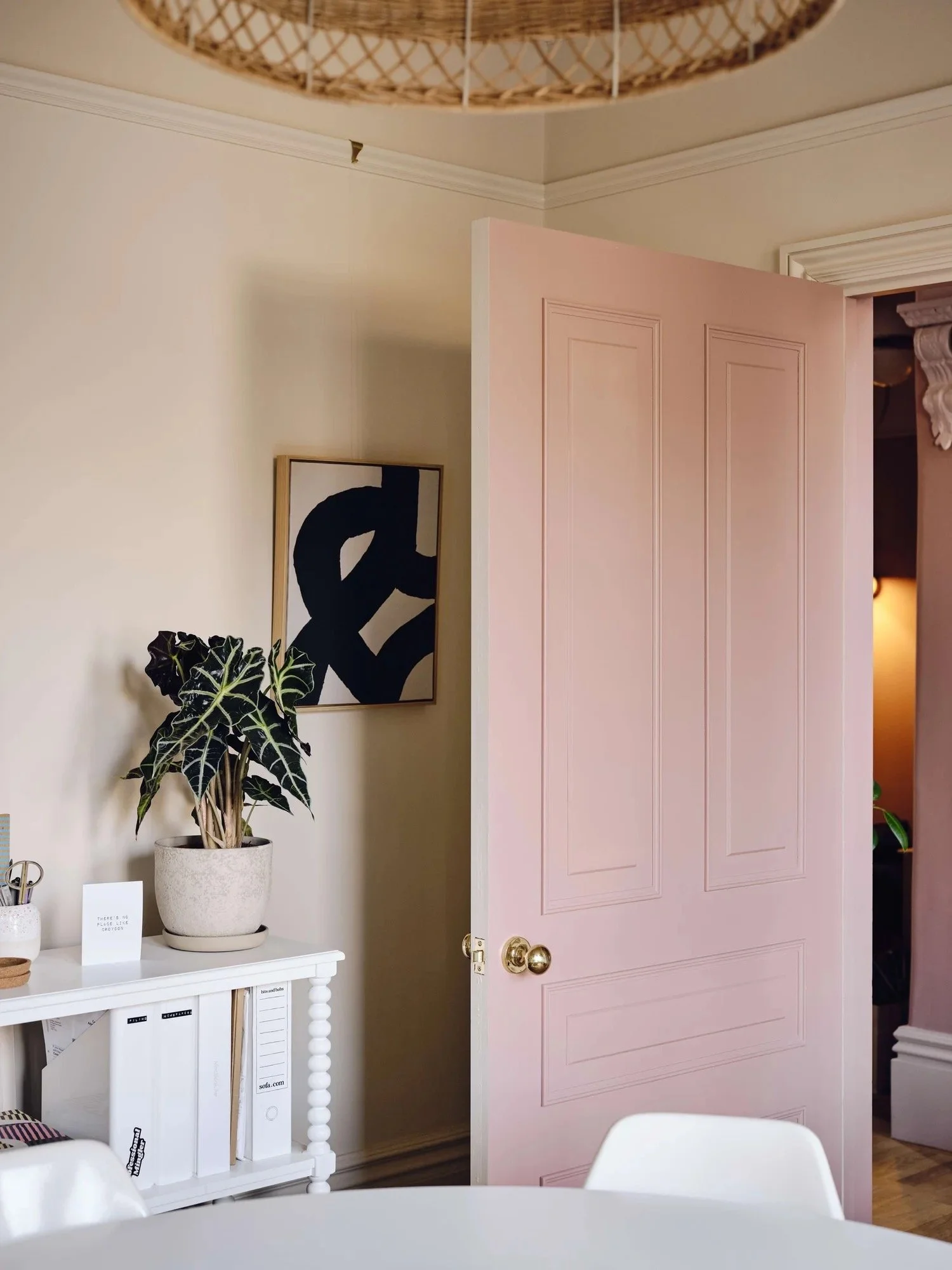 A pink door is partially open in a room with beige walls. A framed black and white abstract art piece hangs to the left of the door. A white table with binders and a small card with writing sits below the art, with a potted plant on top.