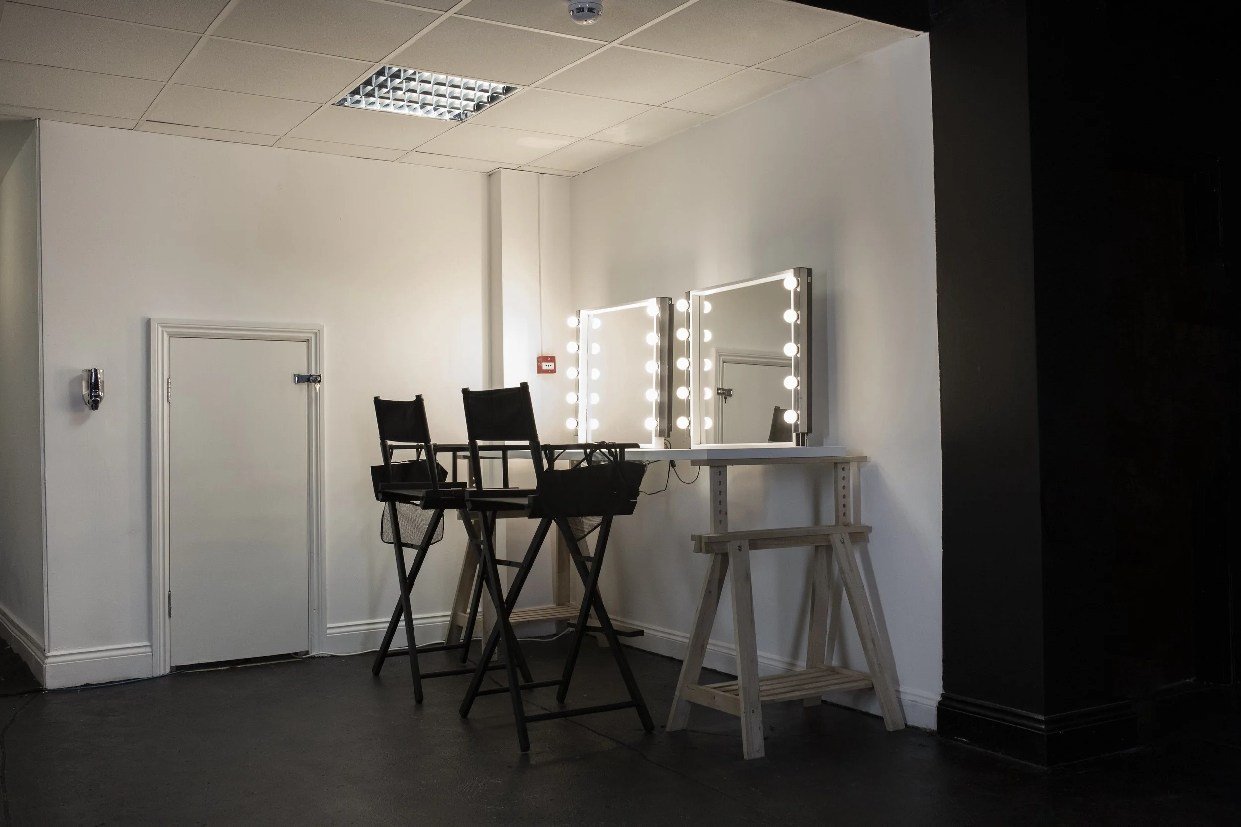 A professional makeup station with two mirrors surrounded by lights, two black makeup chairs, and a small wooden table in a room with white walls and a black wall on the right, with a ceiling light overhead.