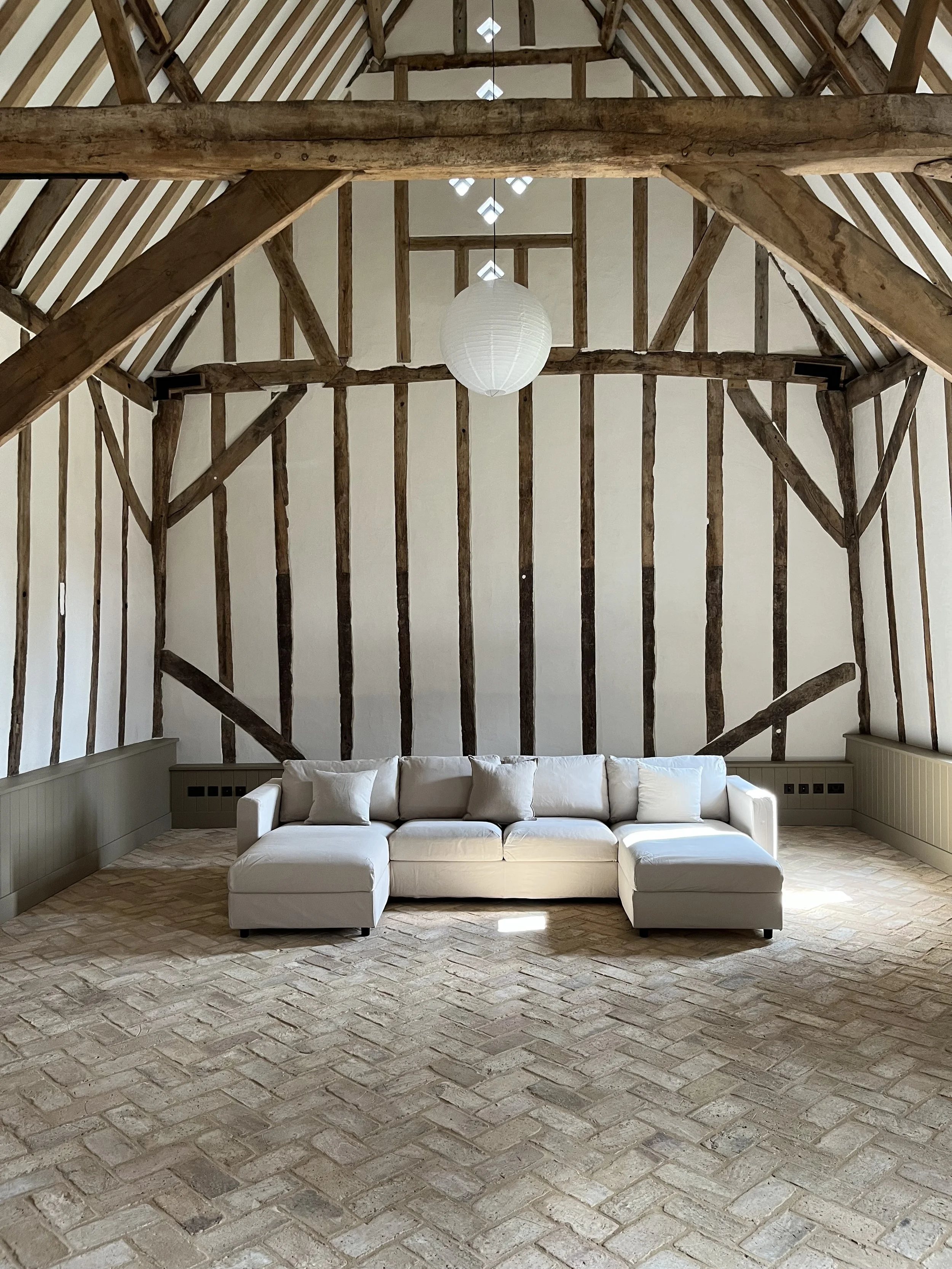 A spacious room with exposed wooden beams, a white sectional sofa with multiple pillows, and a brick floor.
