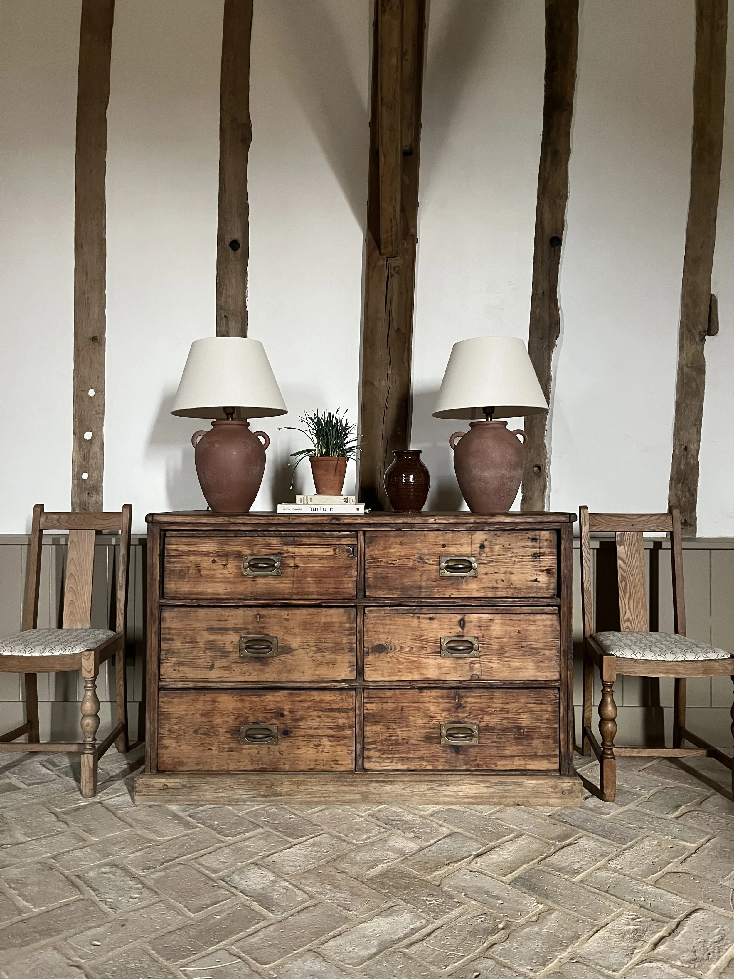 Rustic wooden dresser with eight drawers, adorned with two large beige lamps, a potted plant, and two smaller vases, flanked by two wooden chairs, against a wall with exposed wooden beams.