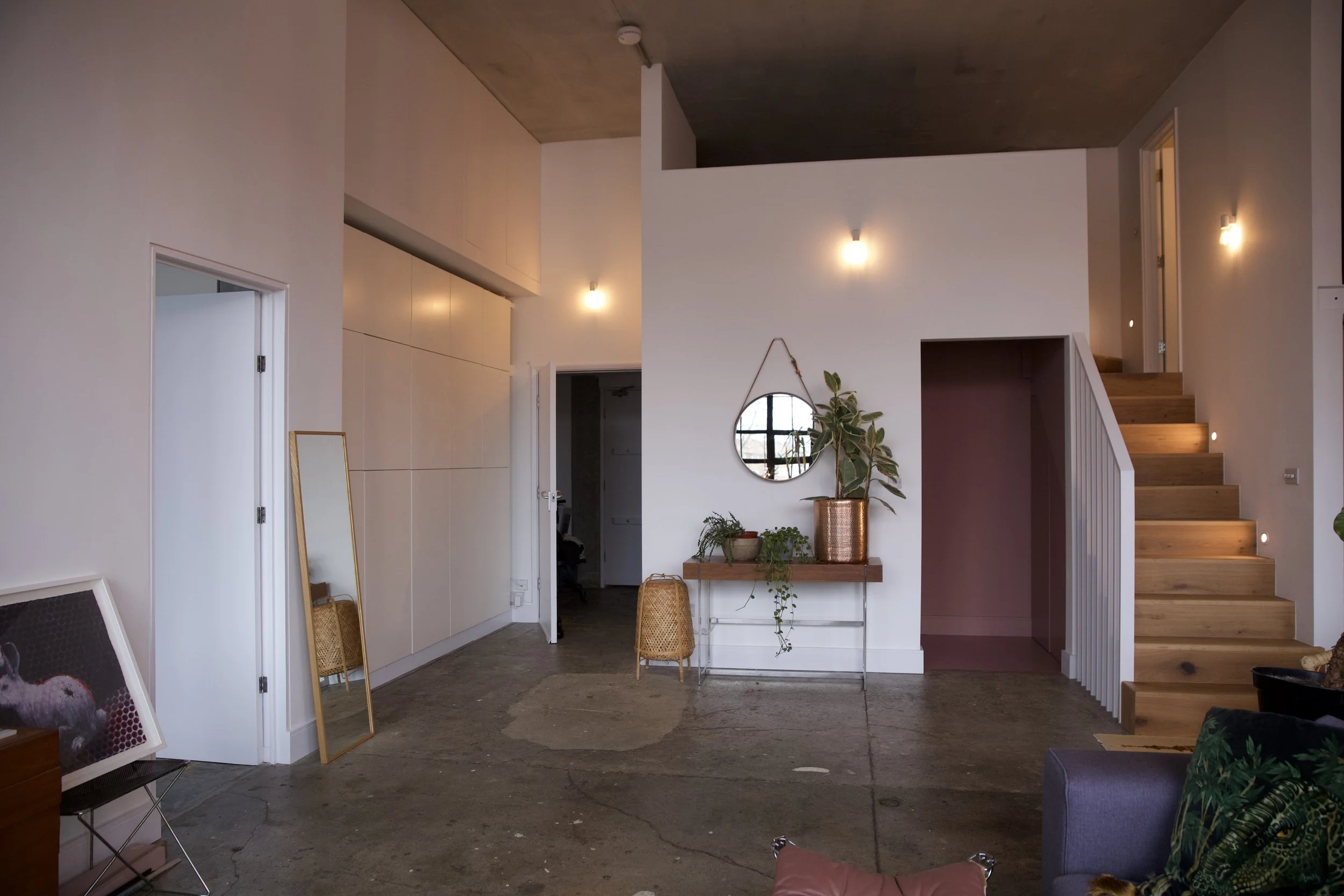 Modern interior of a living space with white walls, wooden stairs, a small wooden console with plants, a mirror, and wall-mounted lights.