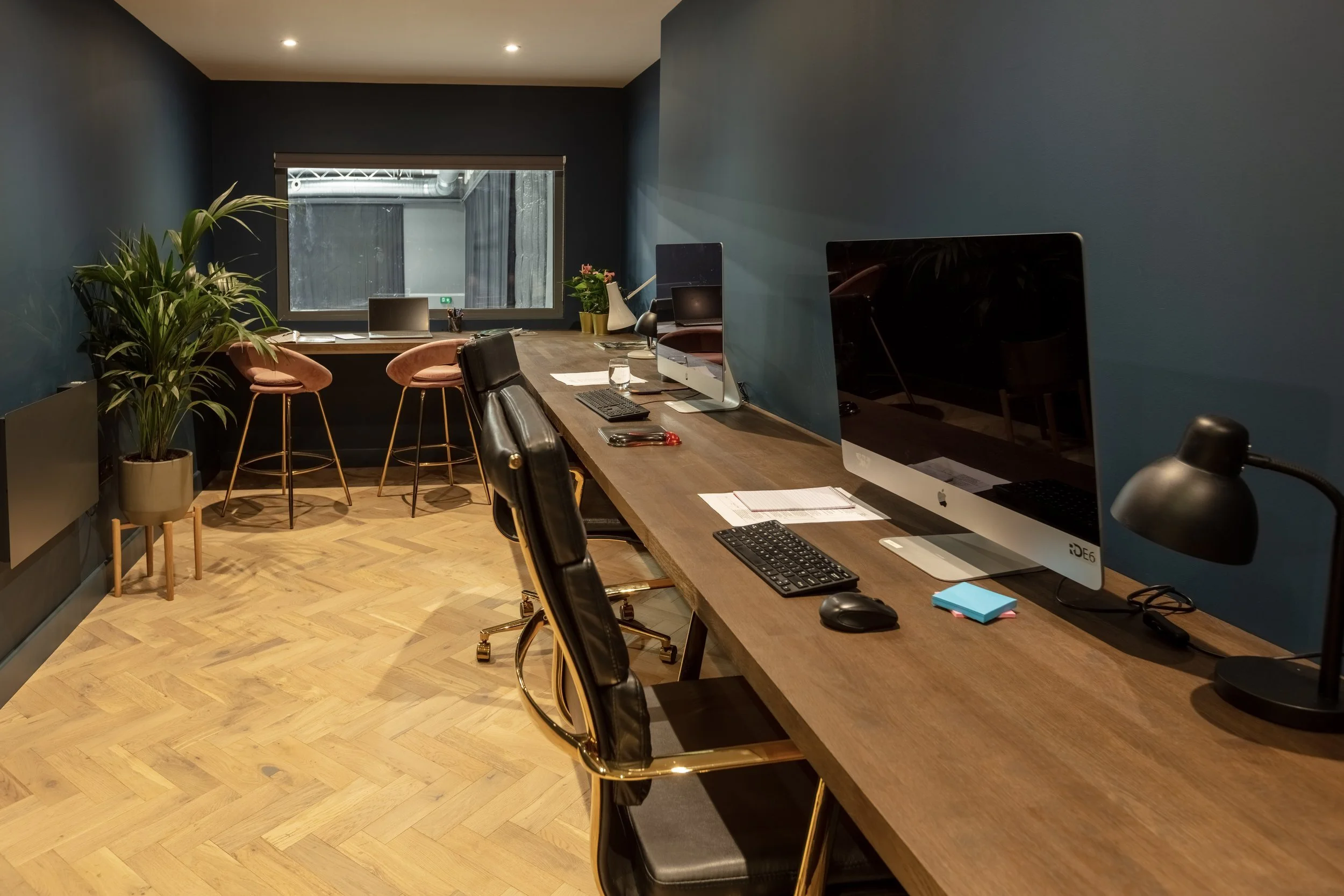 Modern office workspace with a long wooden desk, two large computer monitors, a keyboard, mouse, desk lamp, papers, and a glass of water, against a dark blue wall, with a window at the back and a potted plant on the side.
