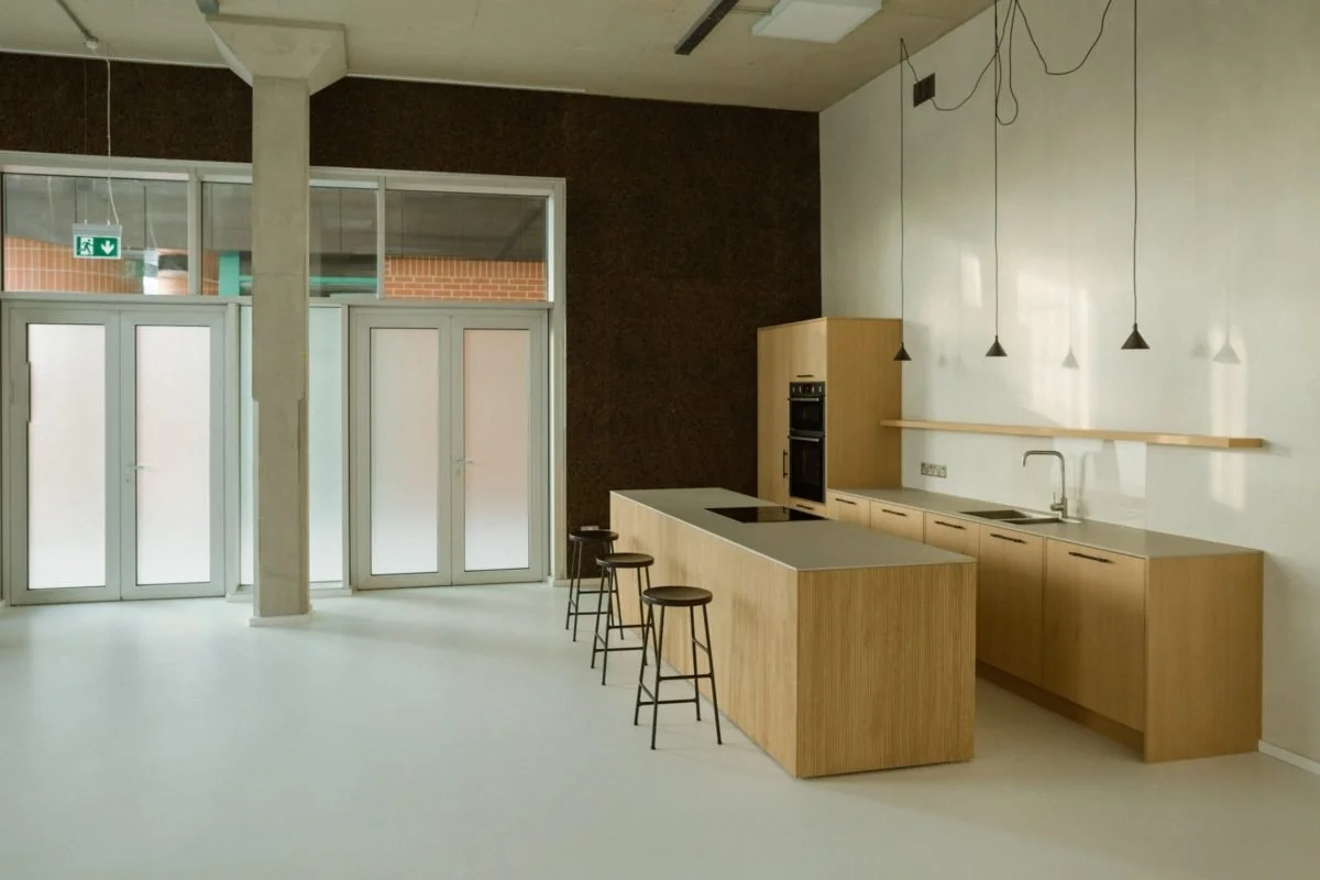 Empty modern kitchen with light wood cabinets, a white countertop, bar stools, pendant lights, and a large windowed entrance.