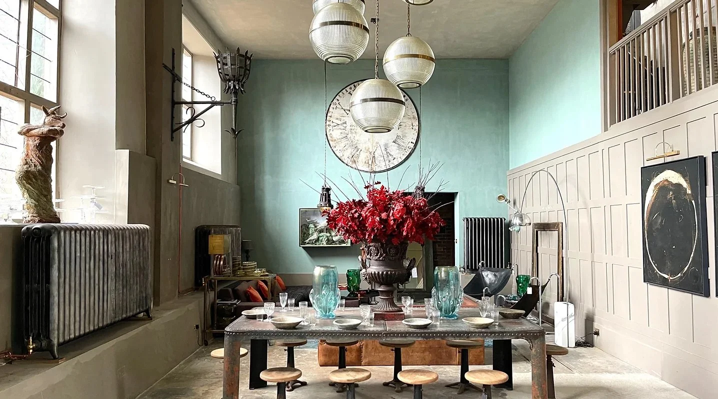 Interior of a stylish dining room with a large vase of red flowers on a long dining table, surrounded by glassware and bowls. The room features high ceilings, large windows, modern hanging light fixtures, wall art, and vintage radiators along the wal
