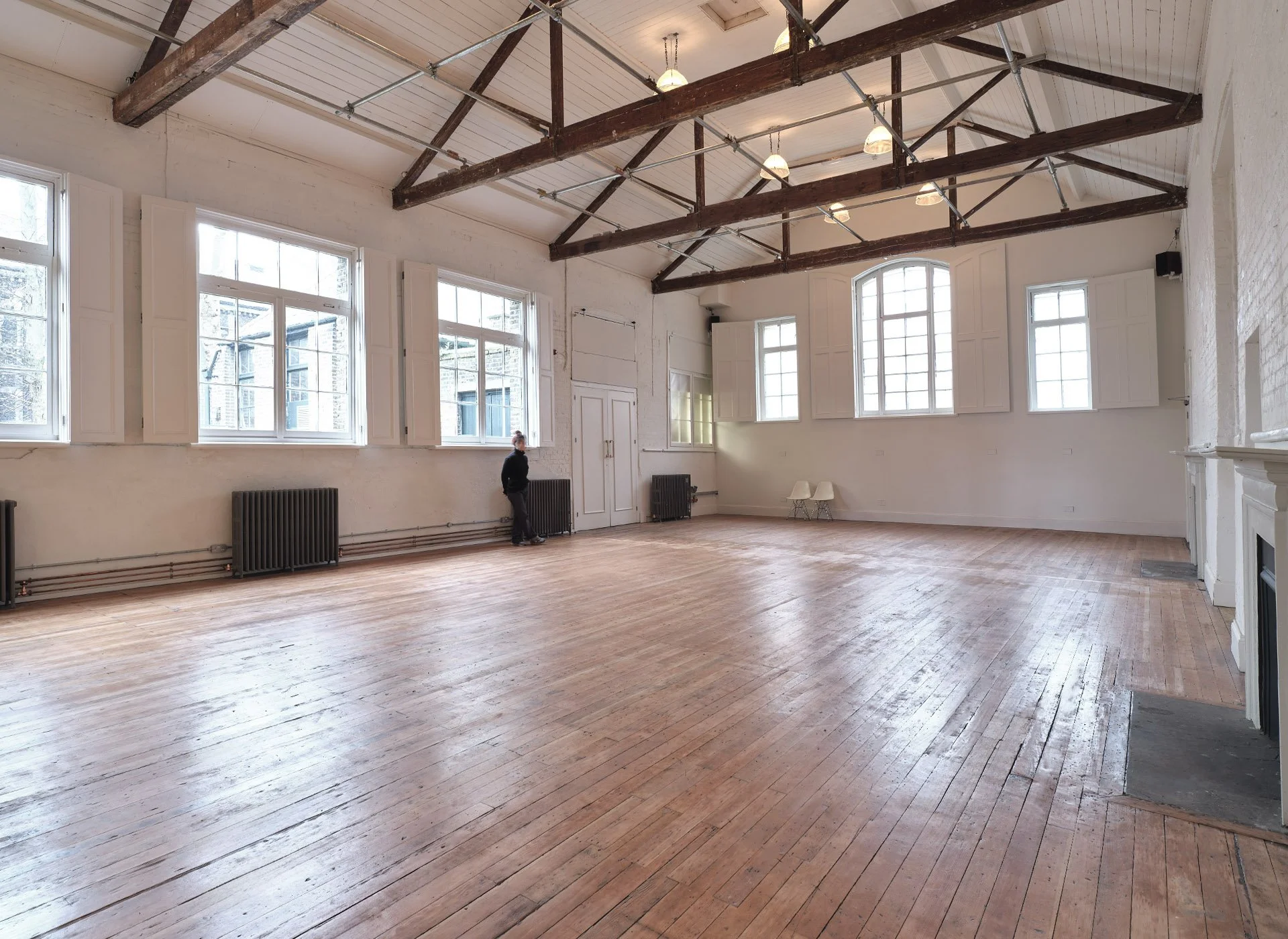 Large, empty room with wooden floors, white walls, multiple large windows with shutters, and exposed wooden beams on the ceiling. A person is standing near the wall on the left side.