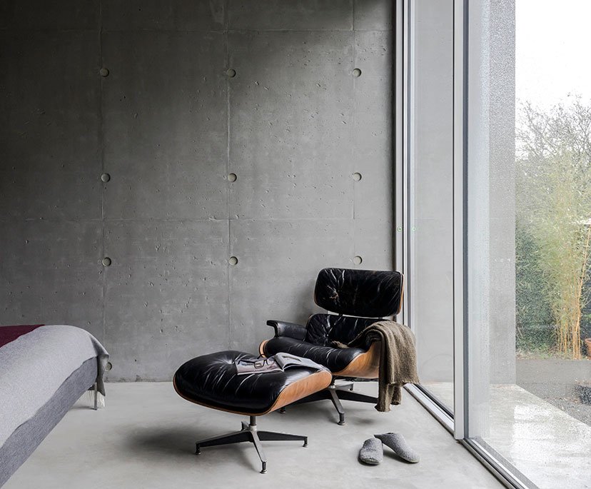 A black leather lounge chair with matching footstool positioned near a large window in a modern minimalist room. A magazine is on the footstool, and a pair of gray slippers are on the light gray concrete floor. The room has a gray textured wall.