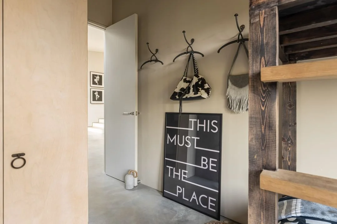 Entryway with abstract black and white sign reading "This must be the place," hanging bags, and wall hooks near a door.