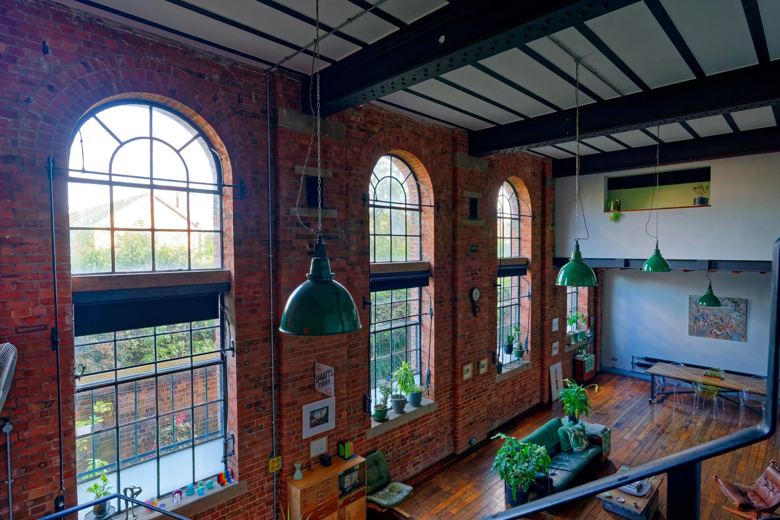 Interior of a spacious loft with brick walls, large arched windows, green pendant lamps, and wooden flooring. Contains a green sofa, potted plants, and a white wall with art and shelves.