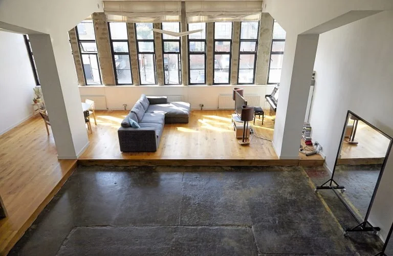 Open living space with large windows, hardwood floors, gray sectional sofa, small dining table with chairs, piano, and standing mirror.