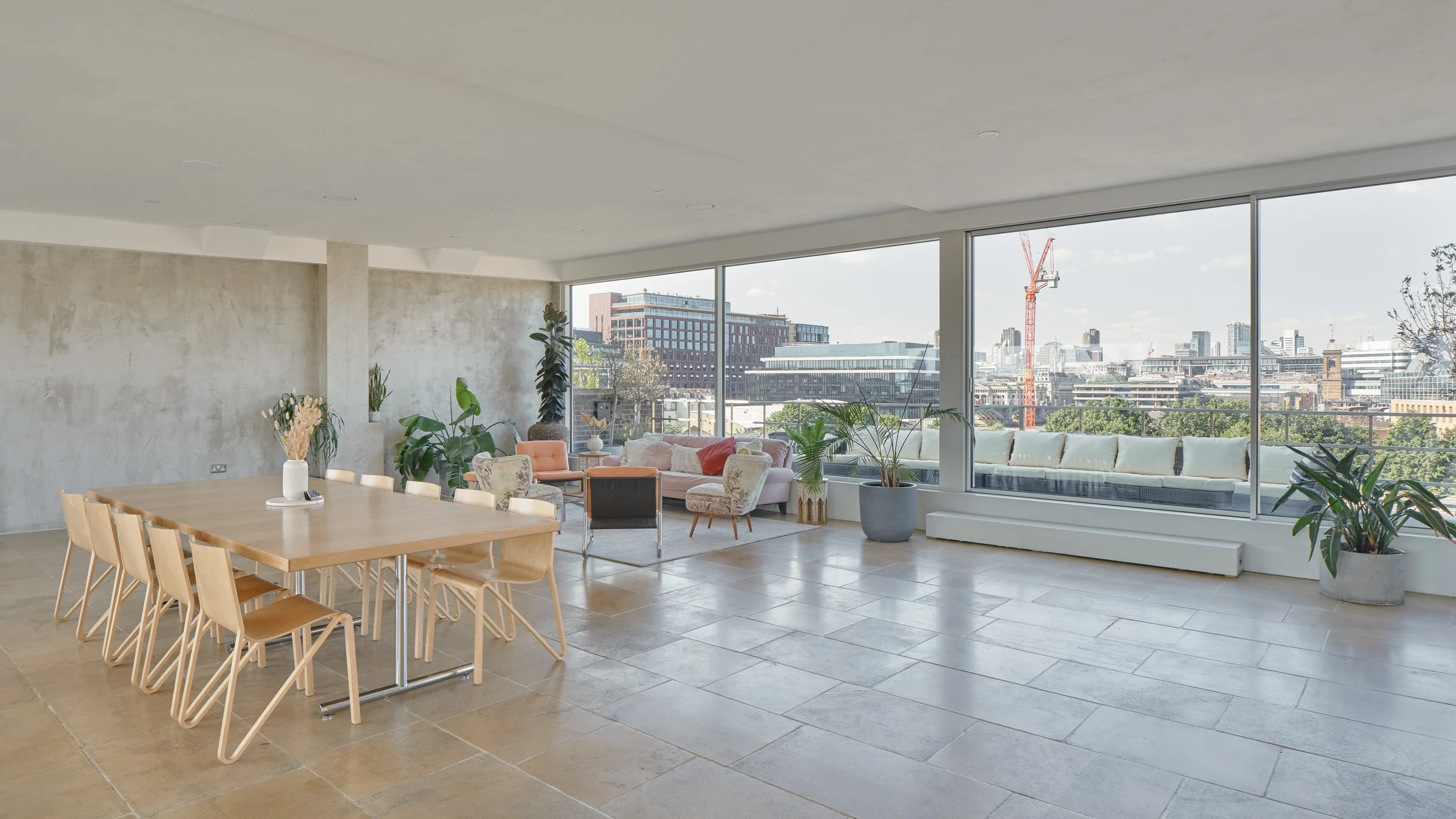 Modern living space with a wooden dining table, multiple chairs, and a seating area with sofas and chairs, large windows with a cityscape view, and potted plants.