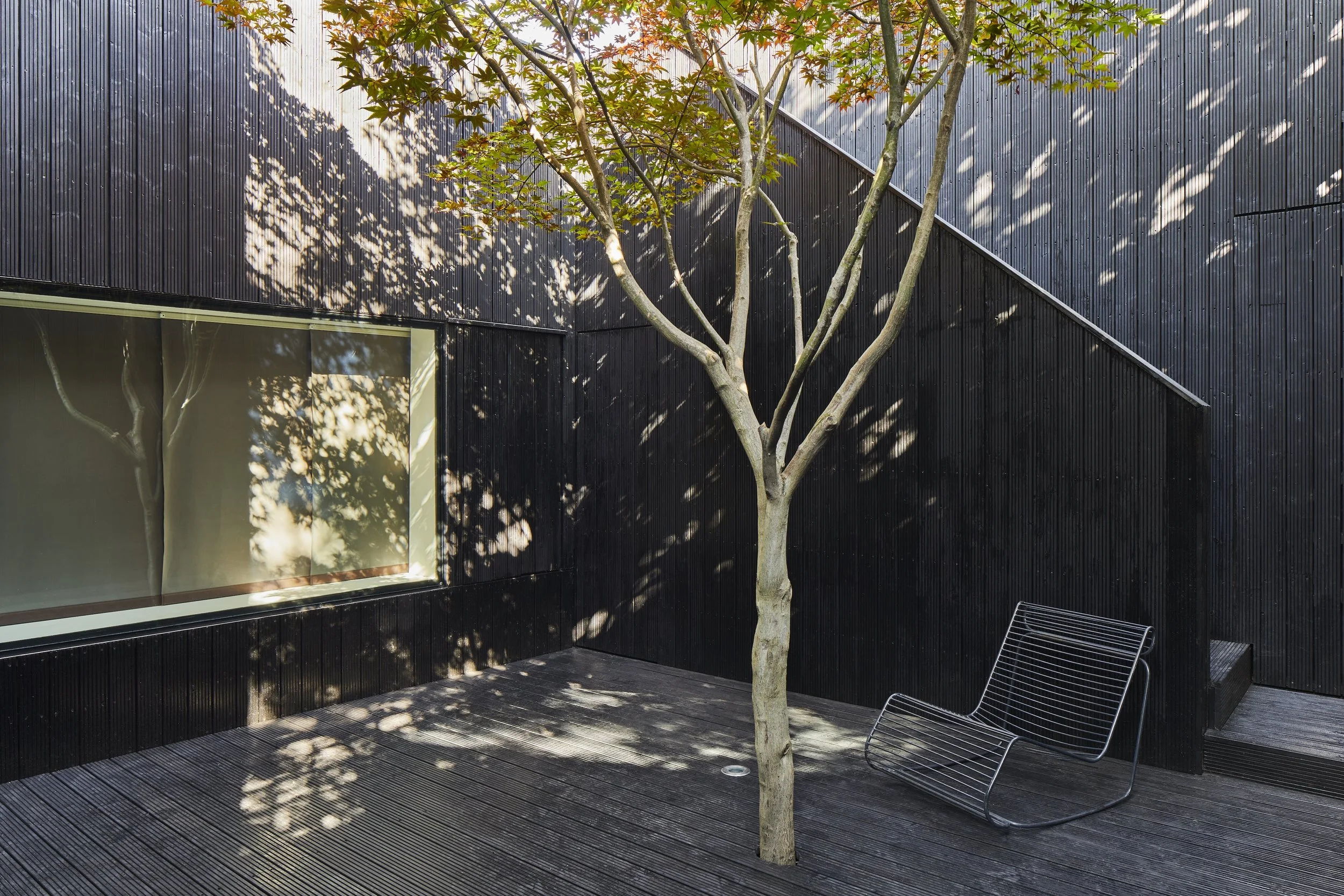 Modern black building exterior with a large window, a tree casting shadows, and a metal chair on a wooden deck.