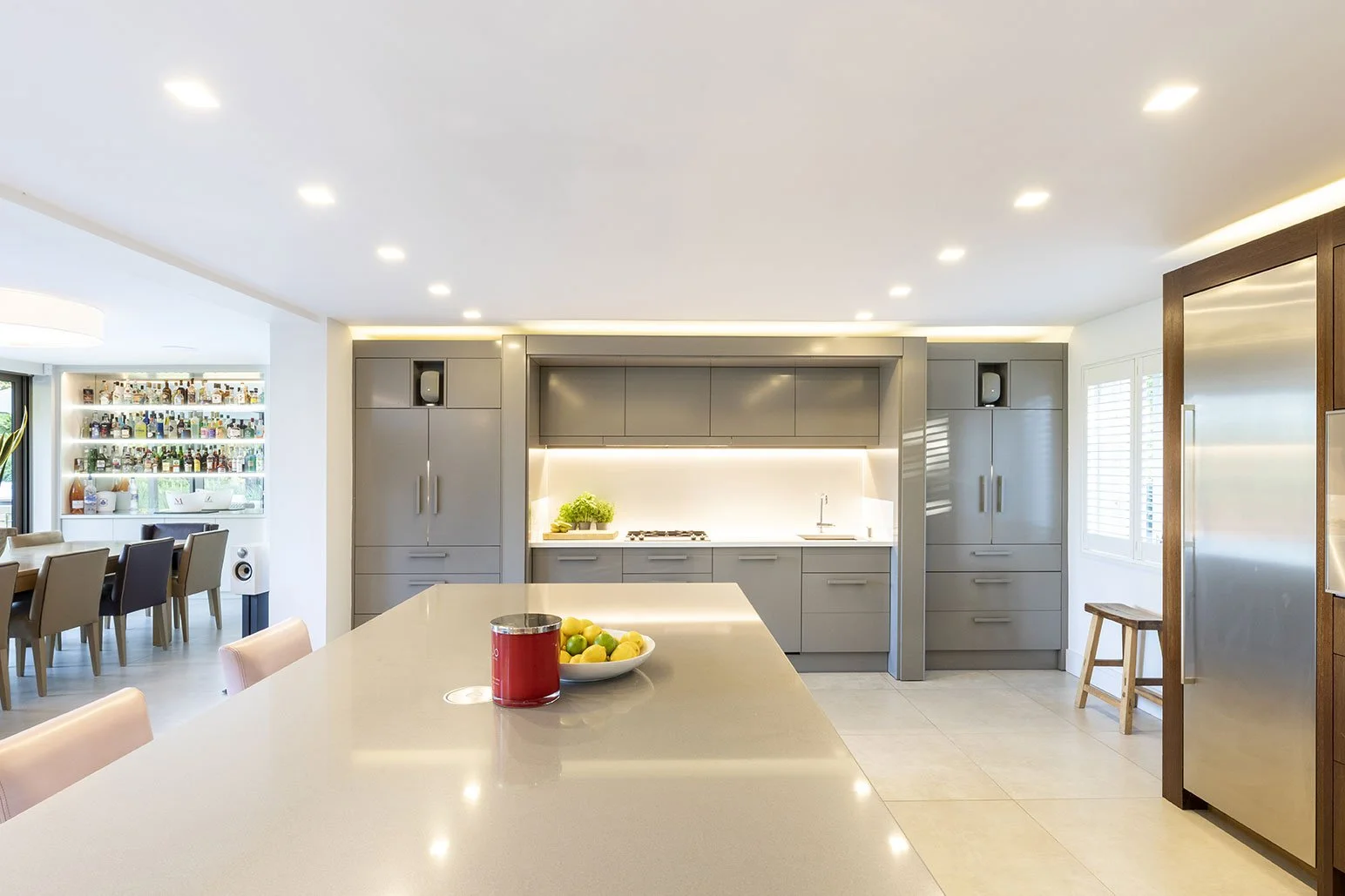 Modern kitchen with gray cabinetry, white countertops, a sink, a stove, built-in speakers, and a bar area with chairs. Adjacent dining area with a large table and chairs, bar shelves with bottles, and a window with shutters.