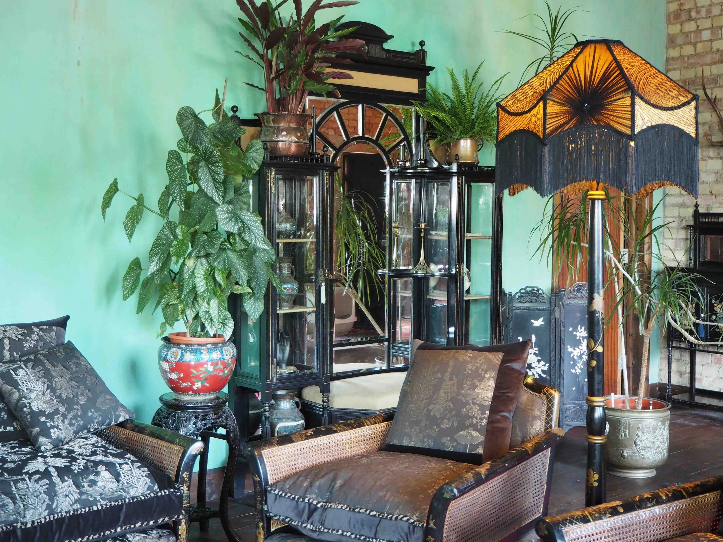 Living room with green wall, vintage black and gold furniture, large potted plants, a standing lamp with an orange and black fringed shade, and decorative glass cabinet.