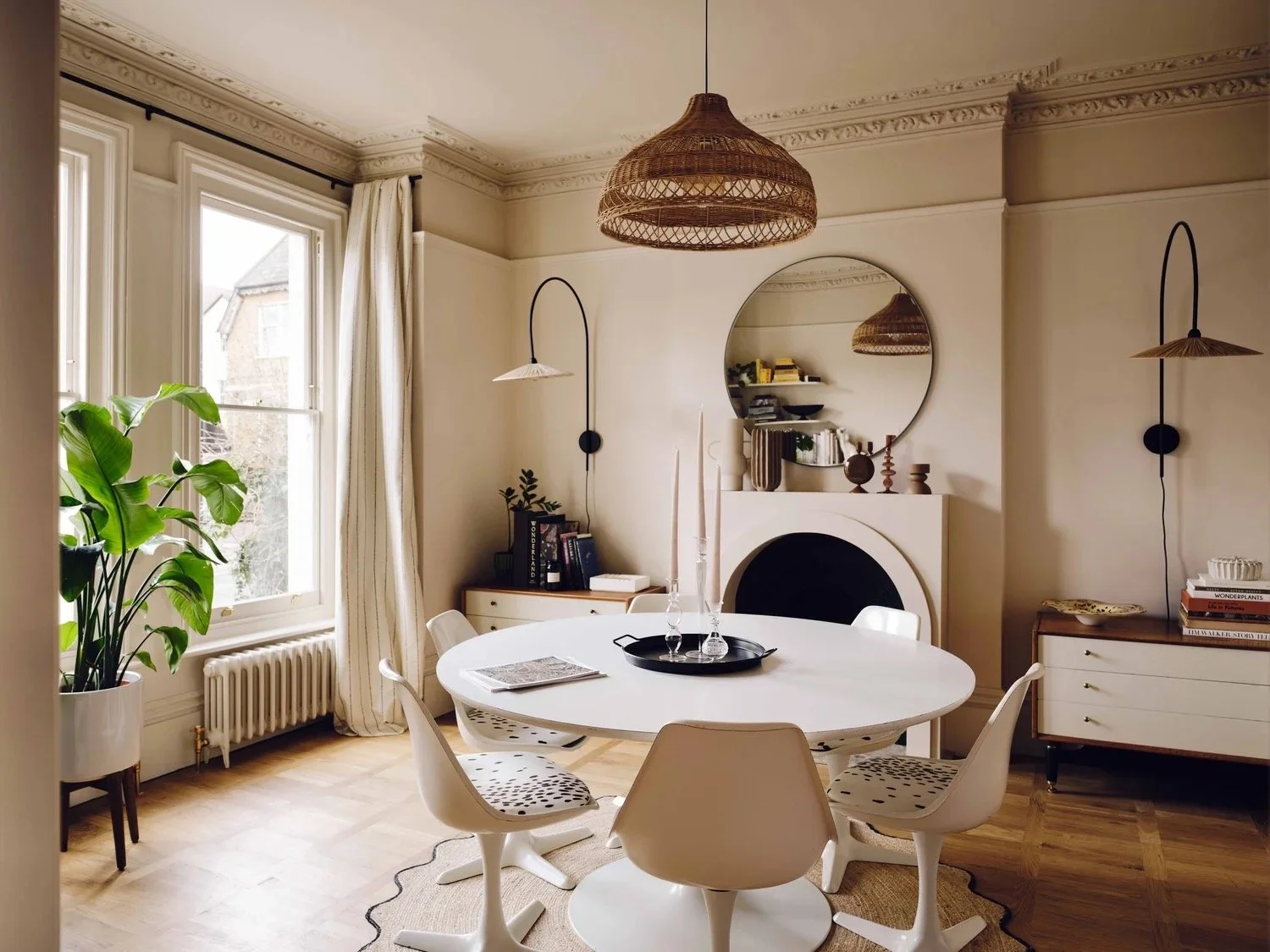 A dining room with a round white table, five modern white chairs, a fireplace with a mirror above it, pendant lights hanging, a sideboard with books, a large green plant near the window, and beige curtains, in a neutral-toned decor.