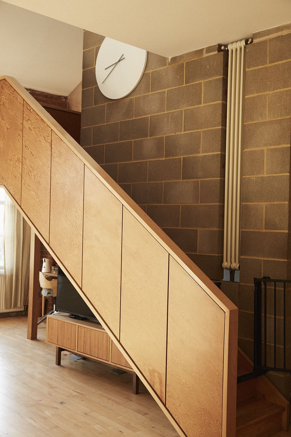 A modern interior with a staircase that has wooden panels on the side, a wall clock, exposed pipes, and a TV stand.