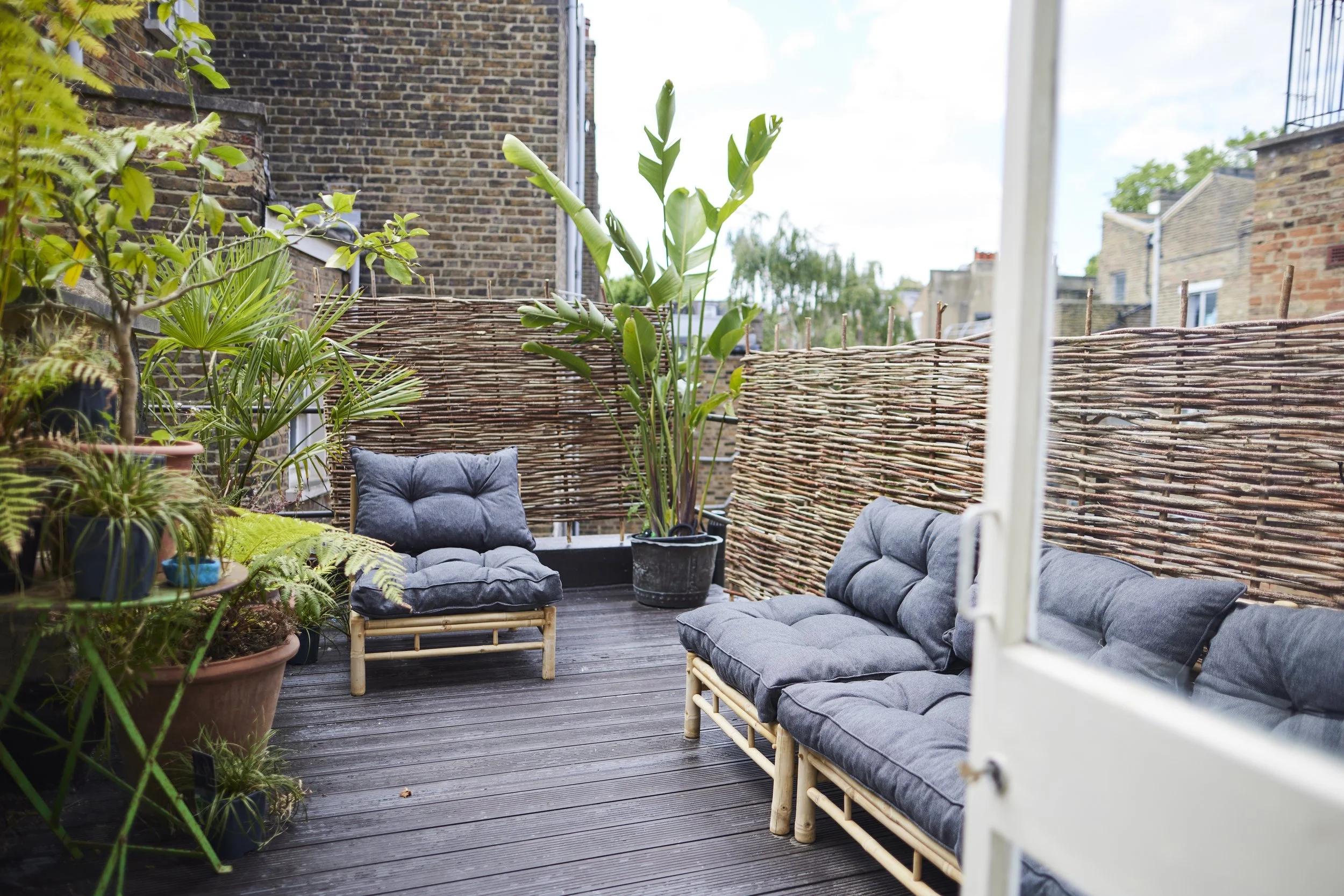 Balcony with potted plants and outdoor furniture, including a cushioned chair and sofa, surrounded by a woven wooden fence, overlooking a cityscape with brick buildings under a partly cloudy sky.