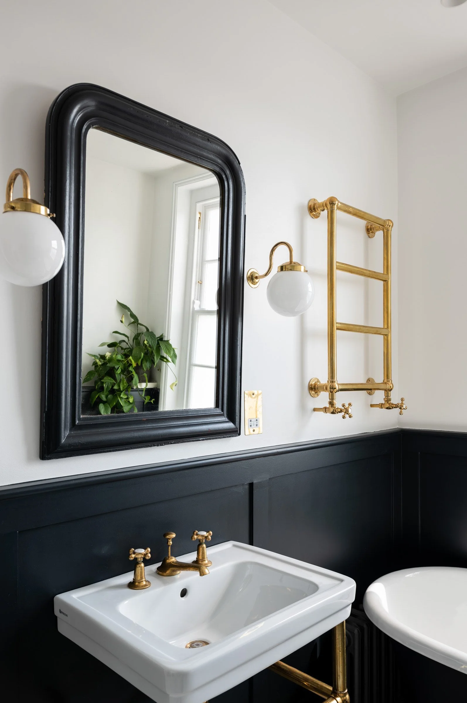 Interior view of a bathroom featuring a rectangular vessel sink with brass fixtures, a large black-framed mirror, gold and brass wall sconces, a gold towel radiator, a white bathtub, and a potted plant near a window.