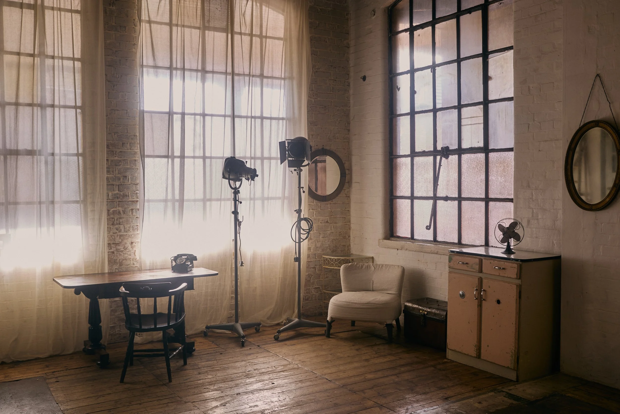 An industrial-style room with large windows, sheer curtains, a small wood table, a black chair, studio lighting equipment, a vintage fan, a beige armchair, a small cabinet, and a mirror on the wall.