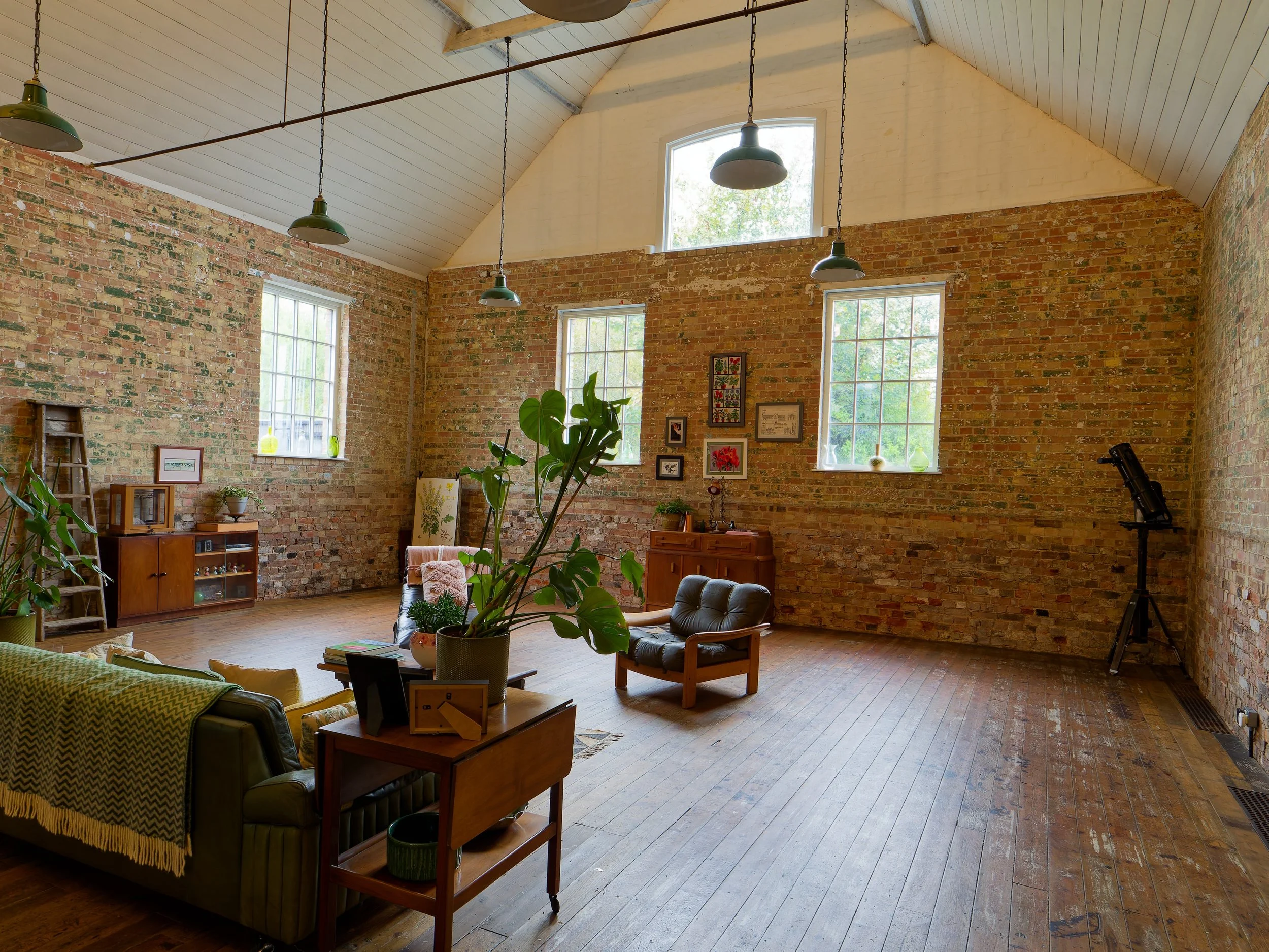 Living room with exposed brick walls, wooden floors, and large windows, decorated with plants, framed pictures, a telescope, and vintage furniture.