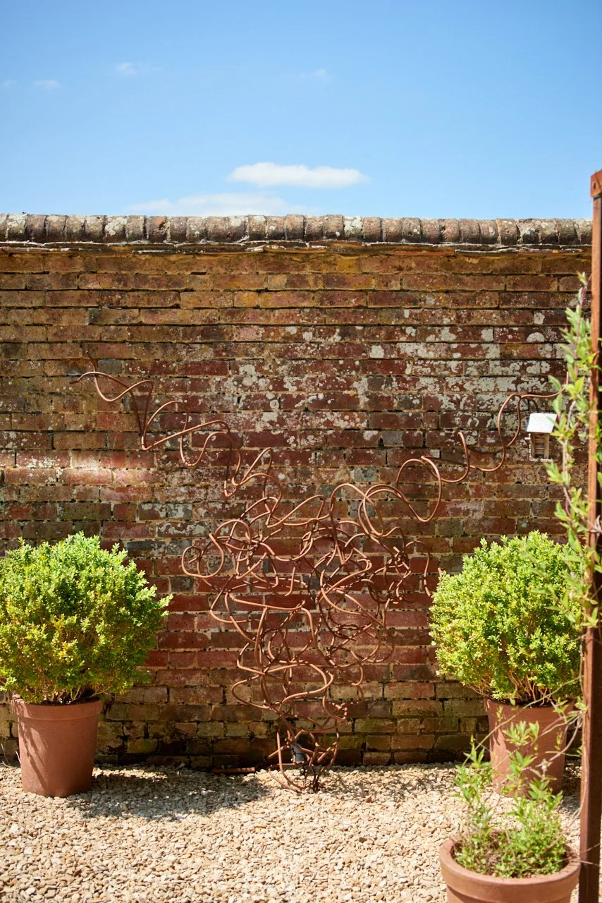 Outdoor scene with a brick wall, two potted green plants, and a metal abstract sculpture in front of the wall, under a blue sky with a few clouds.