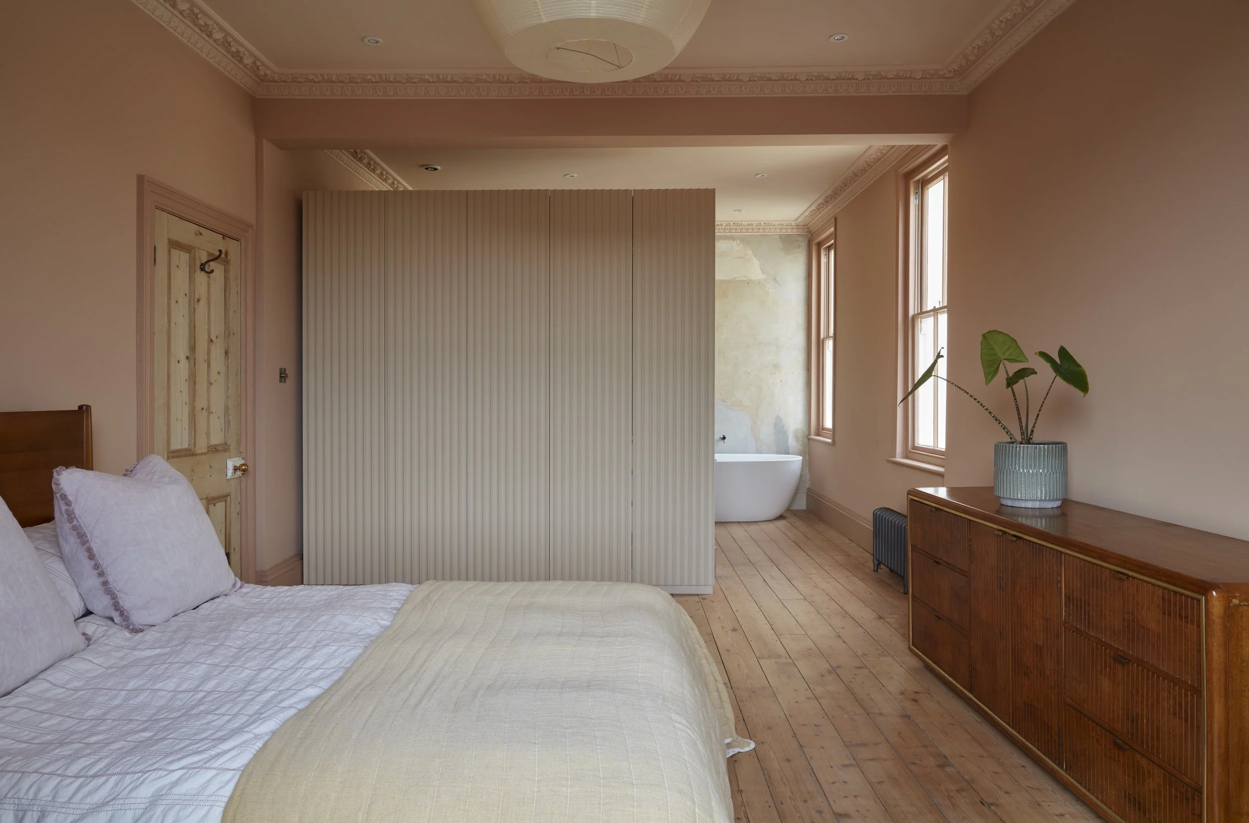 A bedroom with a bed, closet door, wooden dresser with a plant, and a bathtub visible through a partition wall, in a room with beige walls, wooden flooring, and large windows.