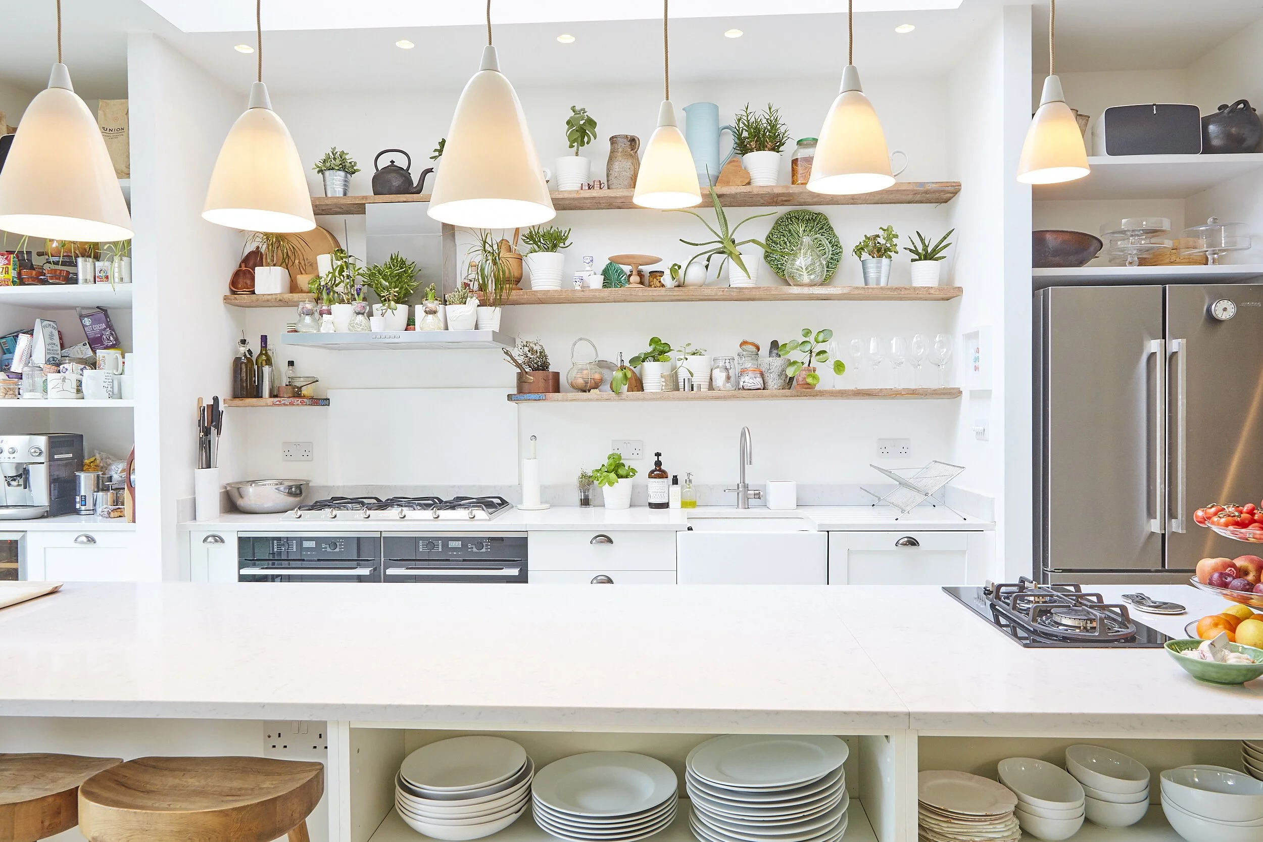 Bright kitchen with white cabinets, open wooden shelves with plants and glassware, pendant lights, stainless steel refrigerator, gas stove, and various kitchen utensils and decorations.