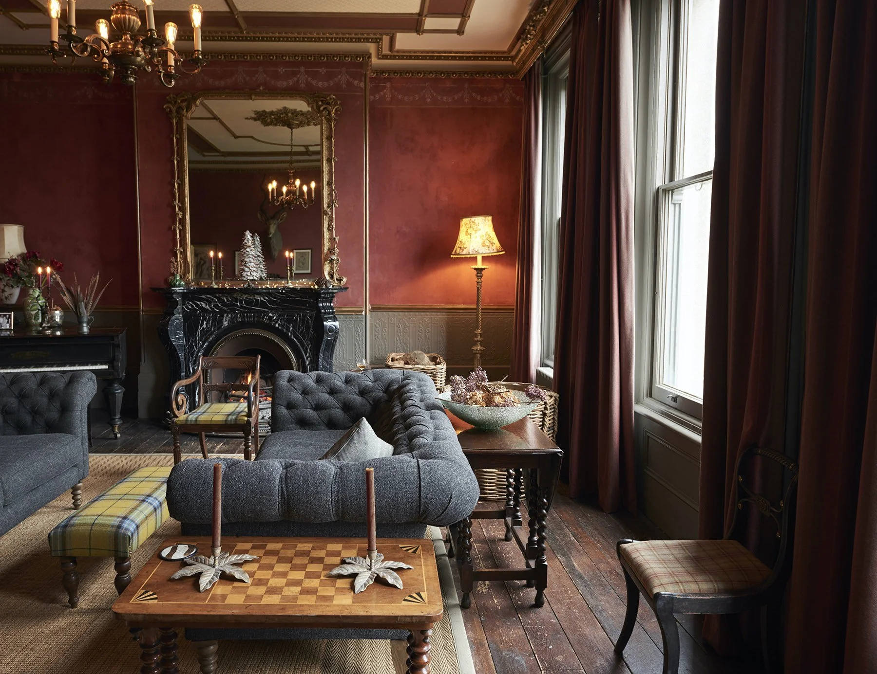 A cozy living room with red walls, wooden flooring, and large windows with heavy curtains. The room features a dark stone fireplace with a large ornate mirror above it, a black piano with a flower arrangement, and a gray tufted sofa. There is a woode