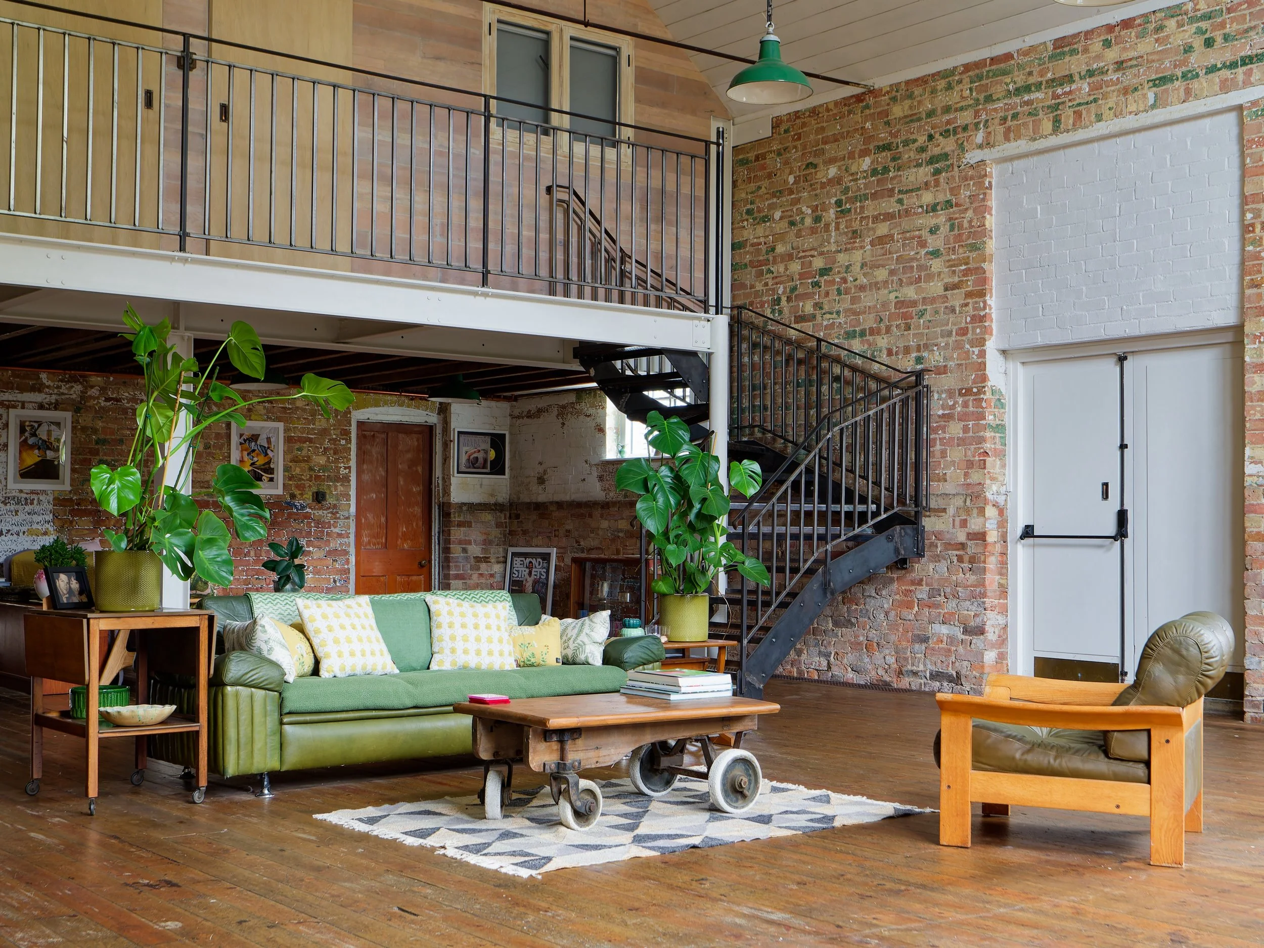 Spacious loft-style living room with exposed brick and wood walls, featuring a green sofa with patterned pillows, a wooden coffee table on wheels, an armchair with a wooden frame, large indoor plants, and a metal spiral staircase leading to a mezzani