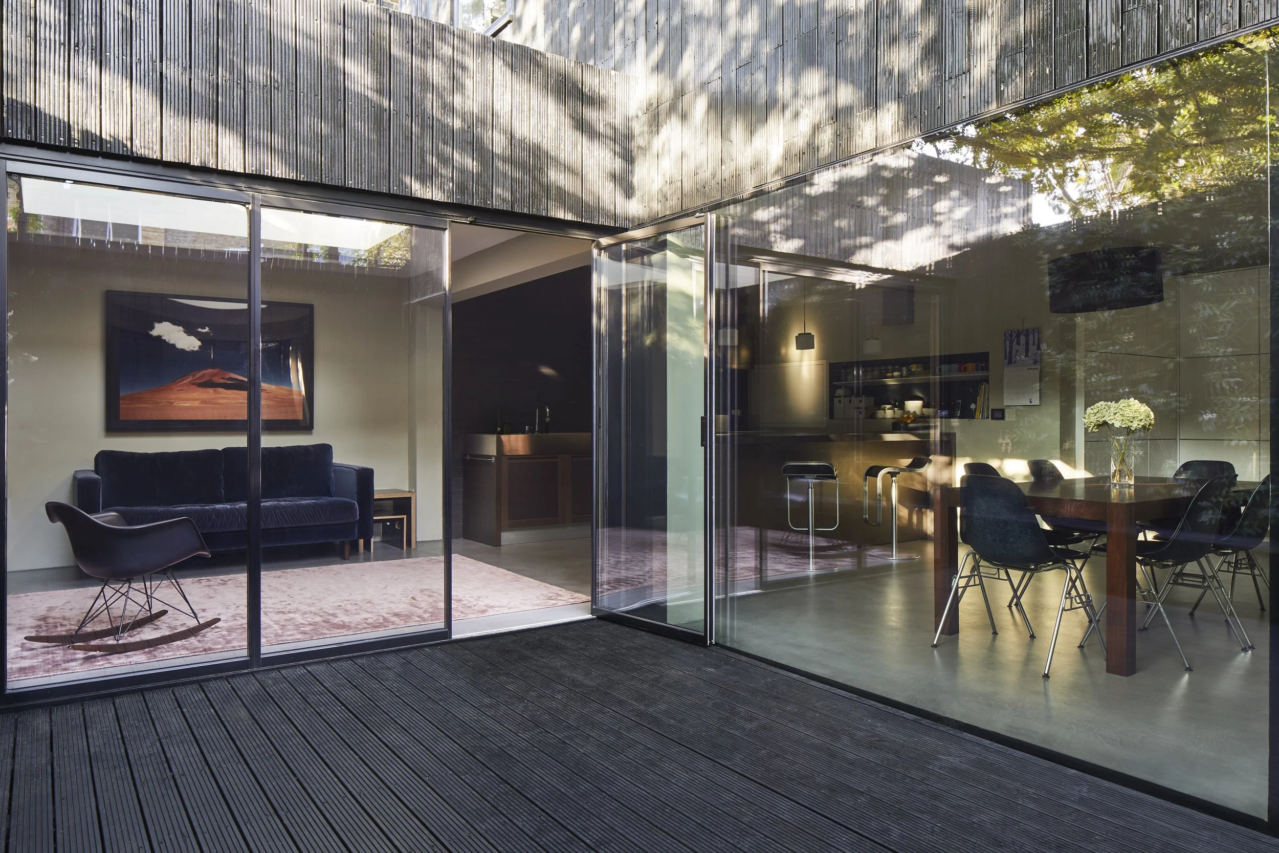 Image of a modern house with glass walls showing a living room and dining area inside. The living room has a dark sofa, a pink rug, and a wall art of a mountain landscape. The dining area has a wooden table, black chairs, and a vase with white flower