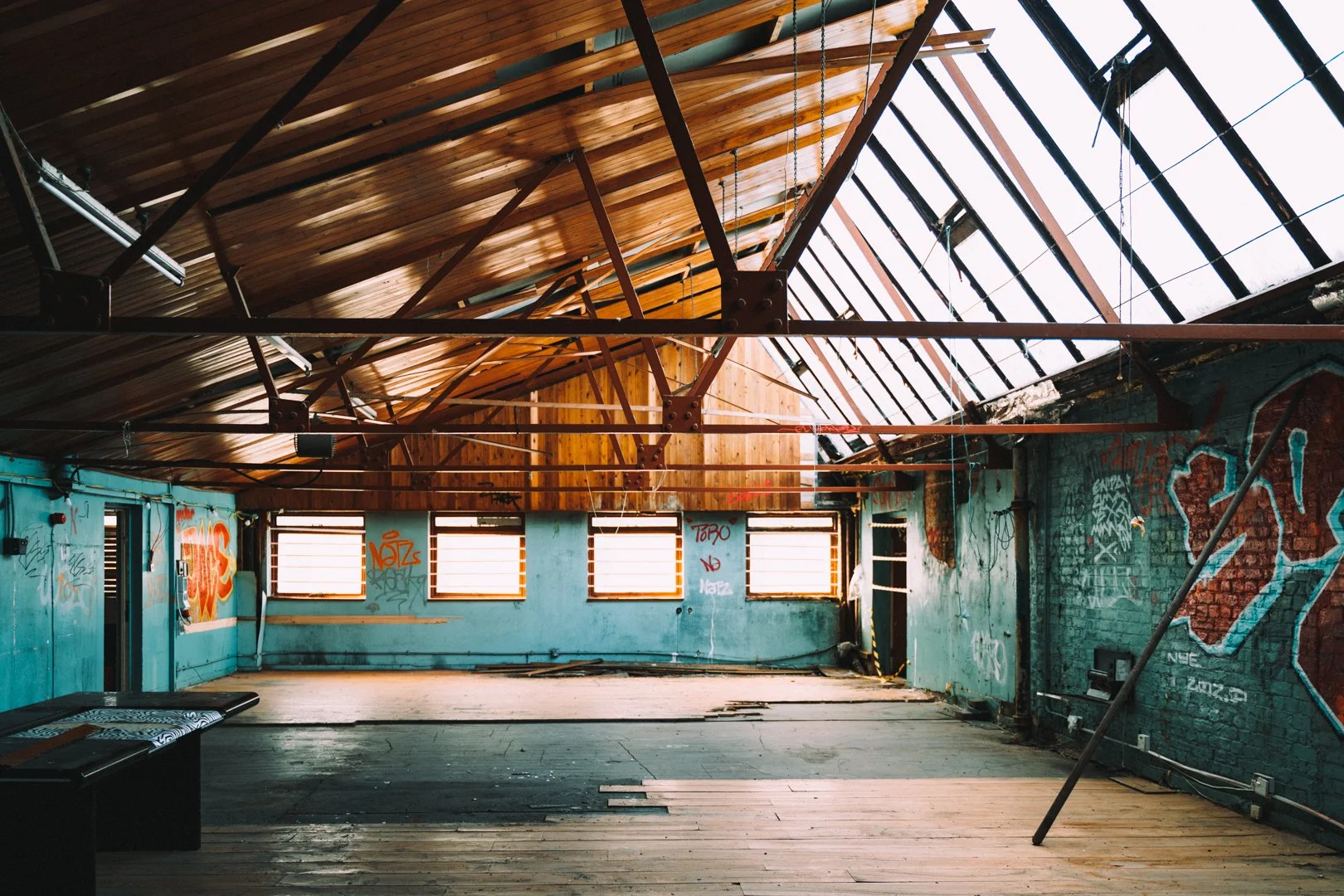 Interior of an abandoned building with graffiti on walls, a wooden ceiling, and broken roof panels allowing natural light inside.