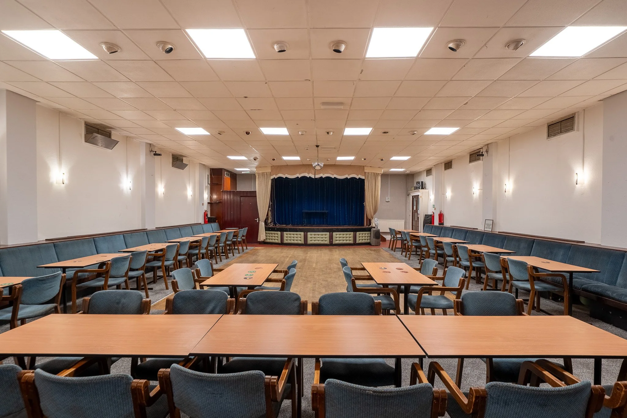 A large, empty community hall with blue upholstered chairs and wooden tables, a stage with a blue curtain, and a ceiling with fluorescent lights.